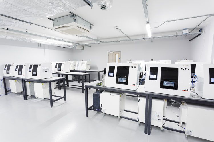 A clean, bright laboratory facility with multiple white and black industrial machines arranged on black tables, featuring overhead LED lighting and mounted ceiling equipment in a sterile white environment.