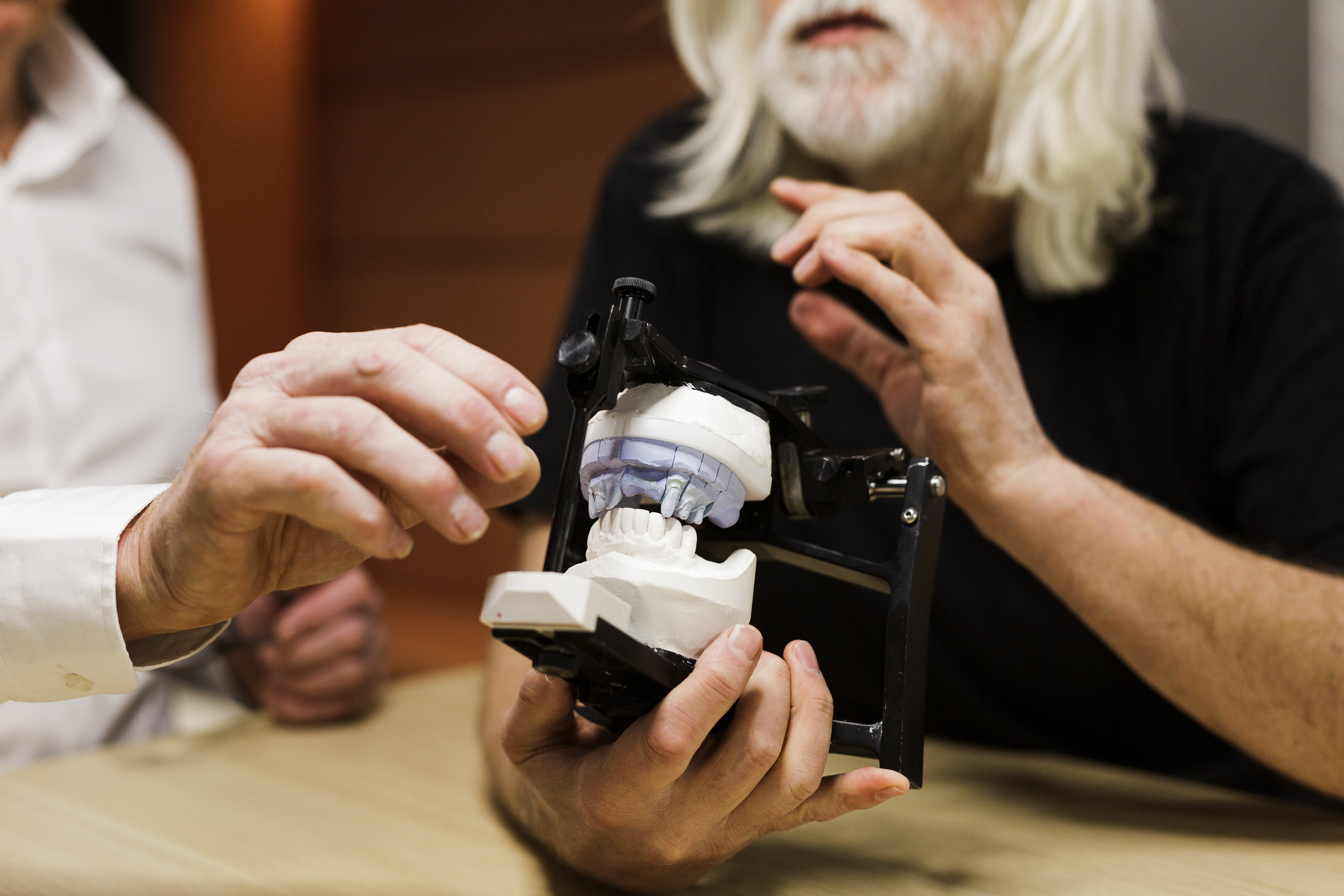 Two dental professionals examine a dental articulator holding white plaster models of teeth with a transparent blue bite registration, demonstrating occlusal analysis in a laboratory setting.