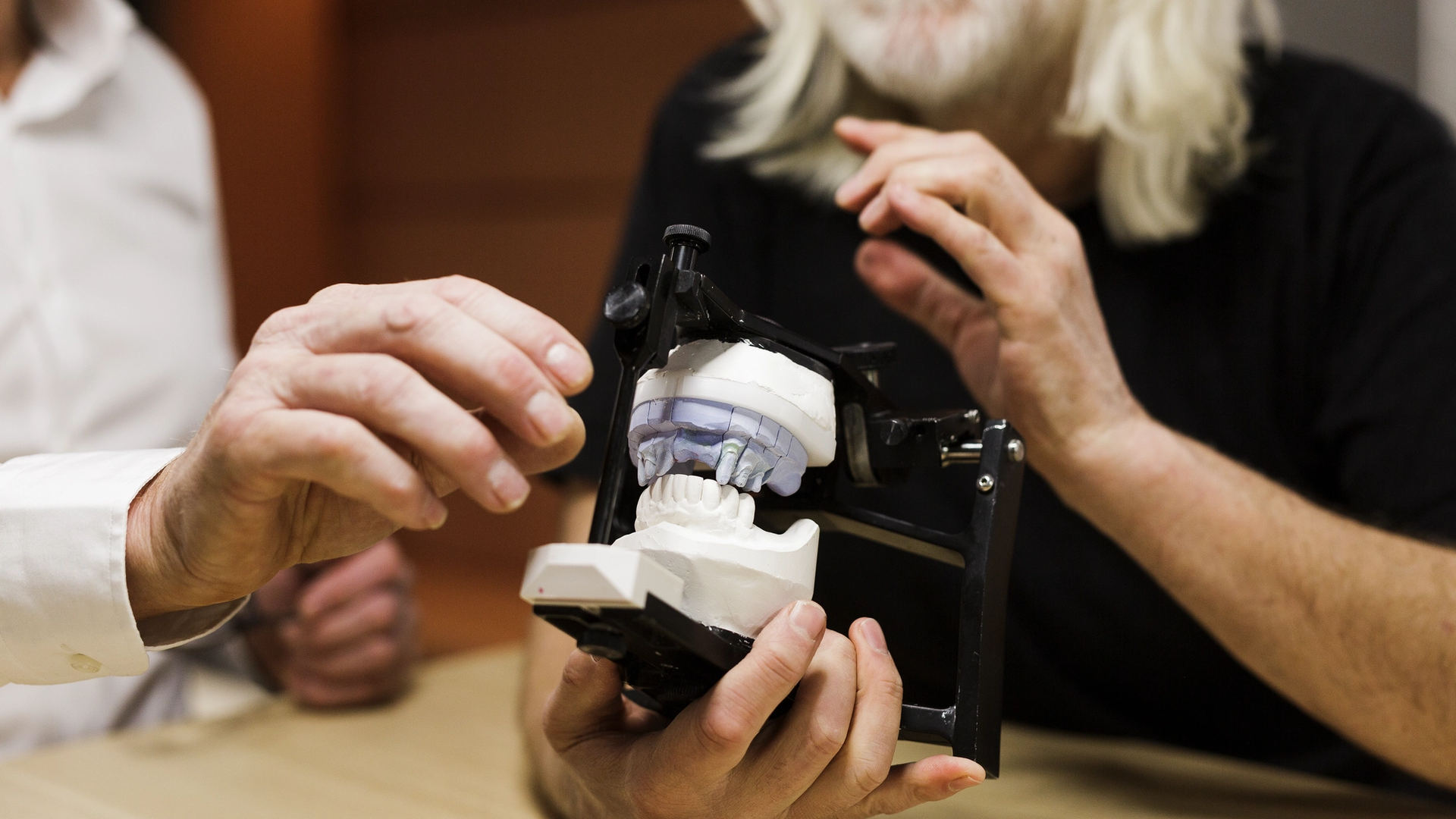 Two dental professionals examine a dental articulator holding white plaster models of teeth with a transparent blue bite registration, demonstrating occlusal analysis in a laboratory setting.