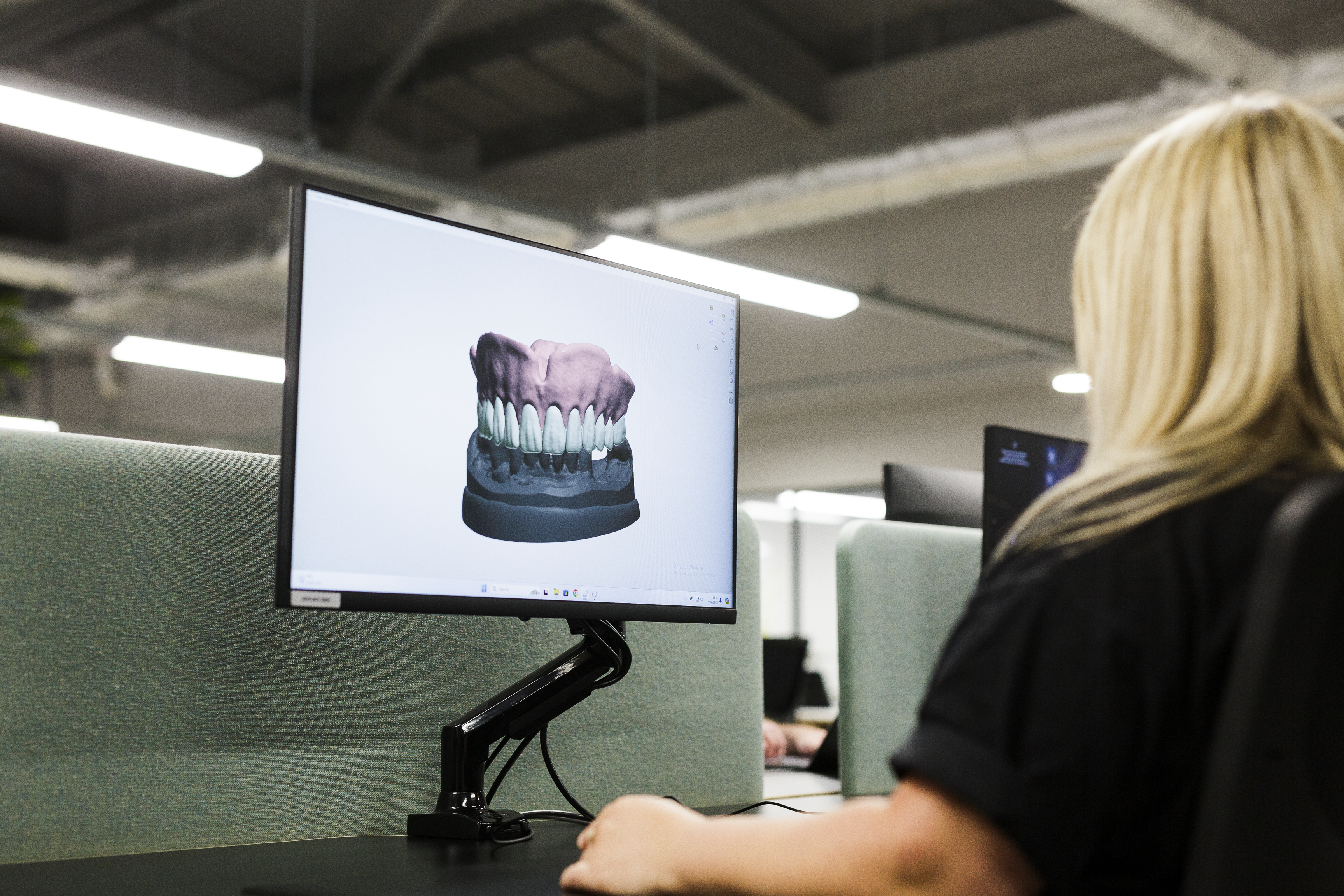 A dental laboratory technician views a 3D digital model of a complete denture arch displayed on a computer monitor, showing upper and lower denture teeth with gum base in a clinical workspace.