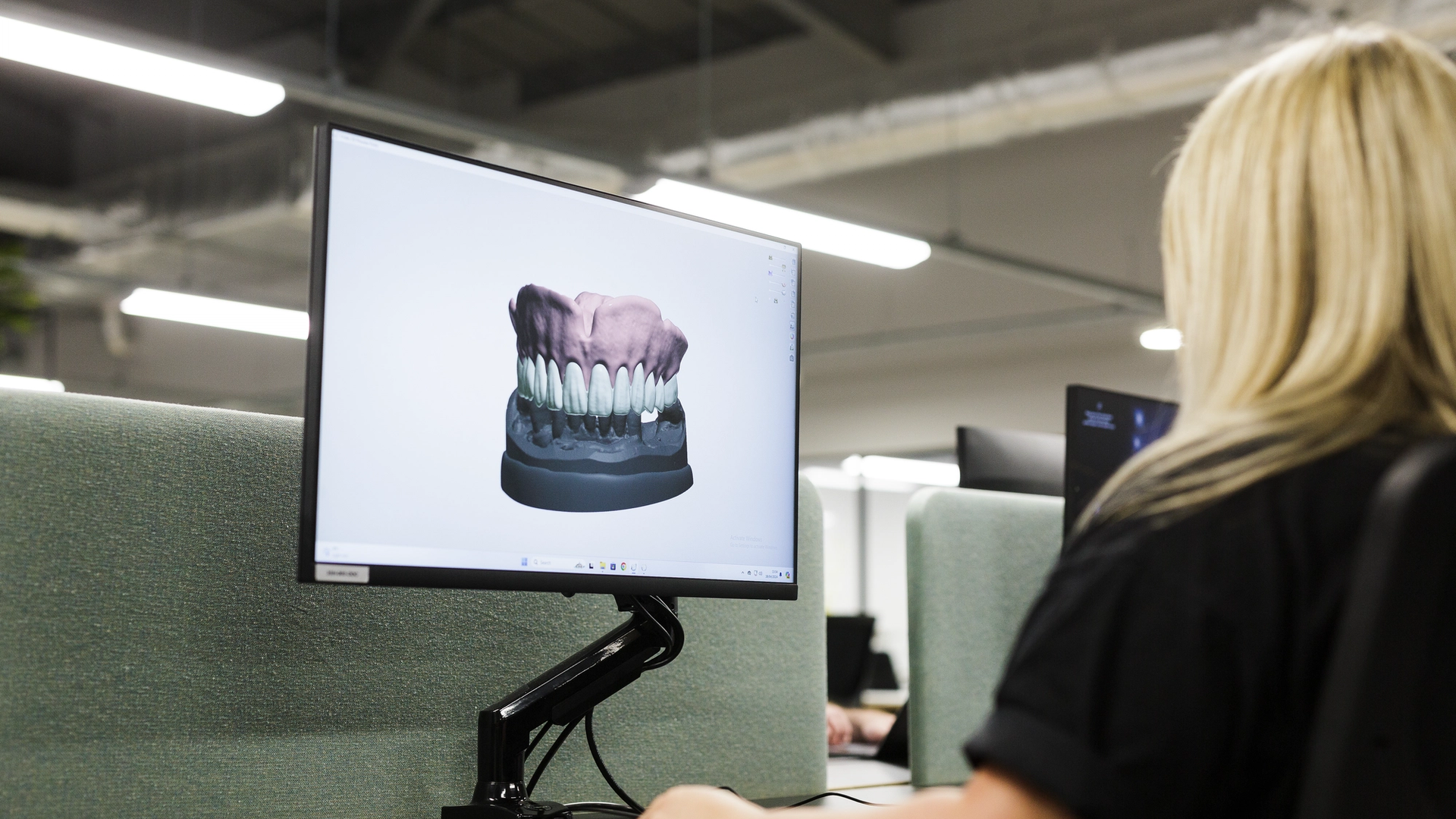 A dental laboratory technician views a 3D digital model of a complete denture arch displayed on a computer monitor, showing upper and lower denture teeth with gum base in a clinical workspace.