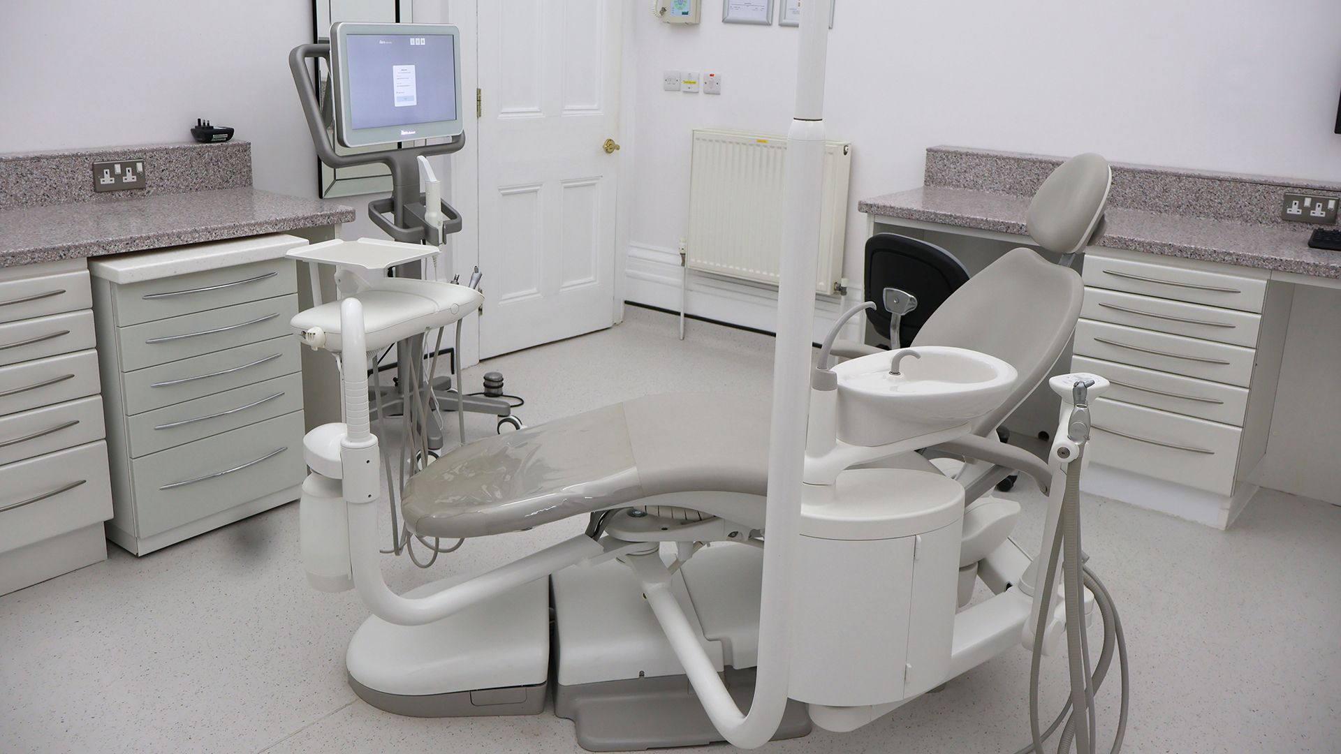 Dental treatment room with two chairs, grey cabinetry, and wall-mounted monitor