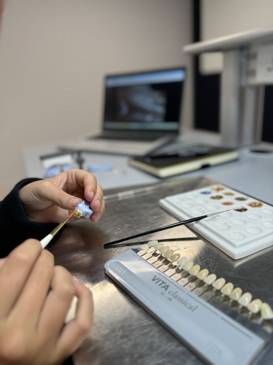 A dental technician uses a brush to apply material to a tooth shade sample while working with VITA color guide tabs and digital imaging equipment in a modern dental laboratory.