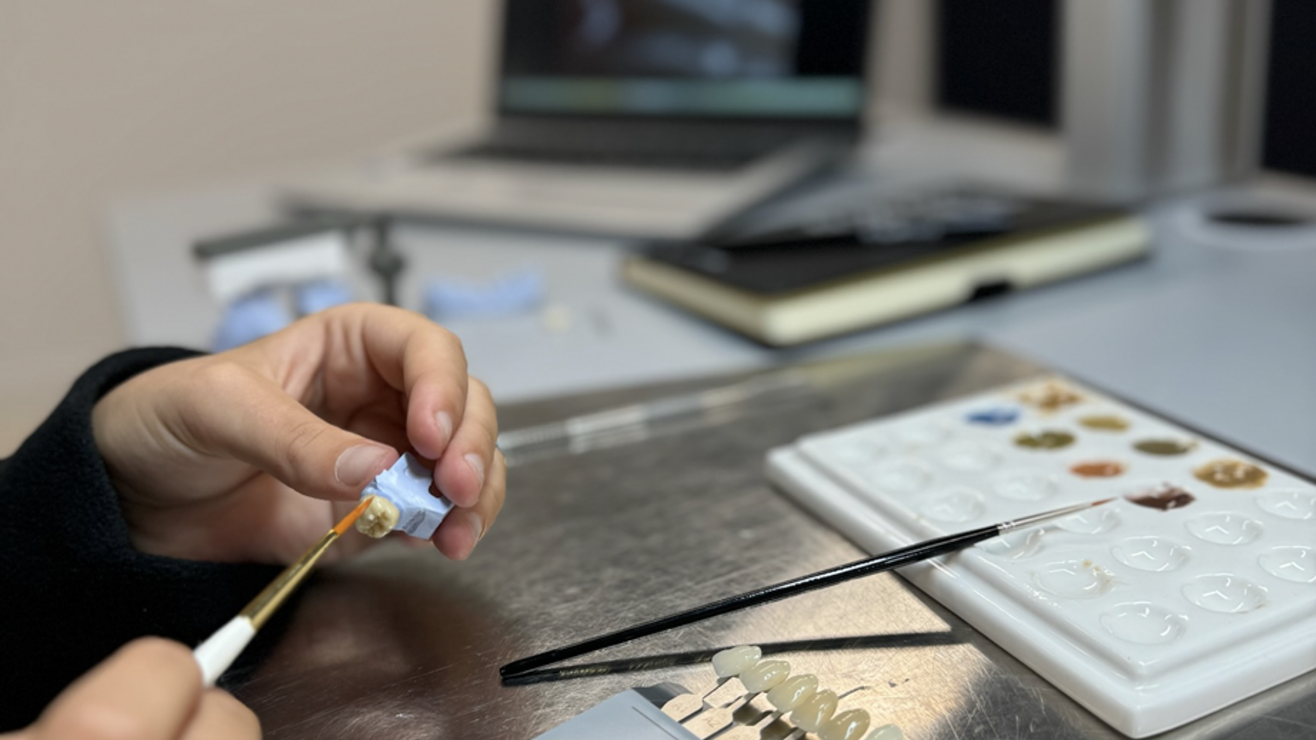 A dental technician uses a brush to apply material to a tooth shade sample while working with VITA color guide tabs and digital imaging equipment in a modern dental laboratory.