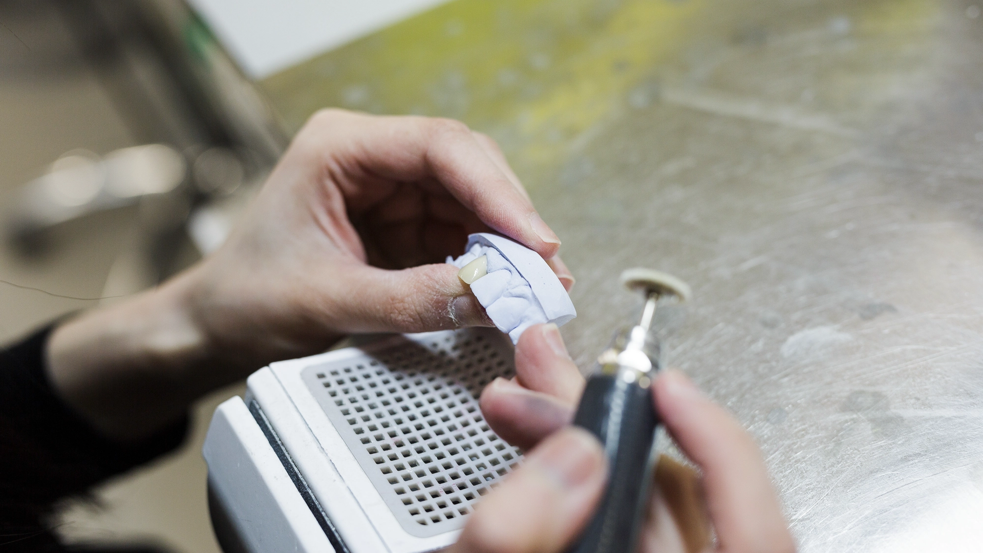 A dental technician holds a milled ceramic dental crown wrapped in protective material while using a rotary instrument to adjust and finish the restoration on a work surface.
