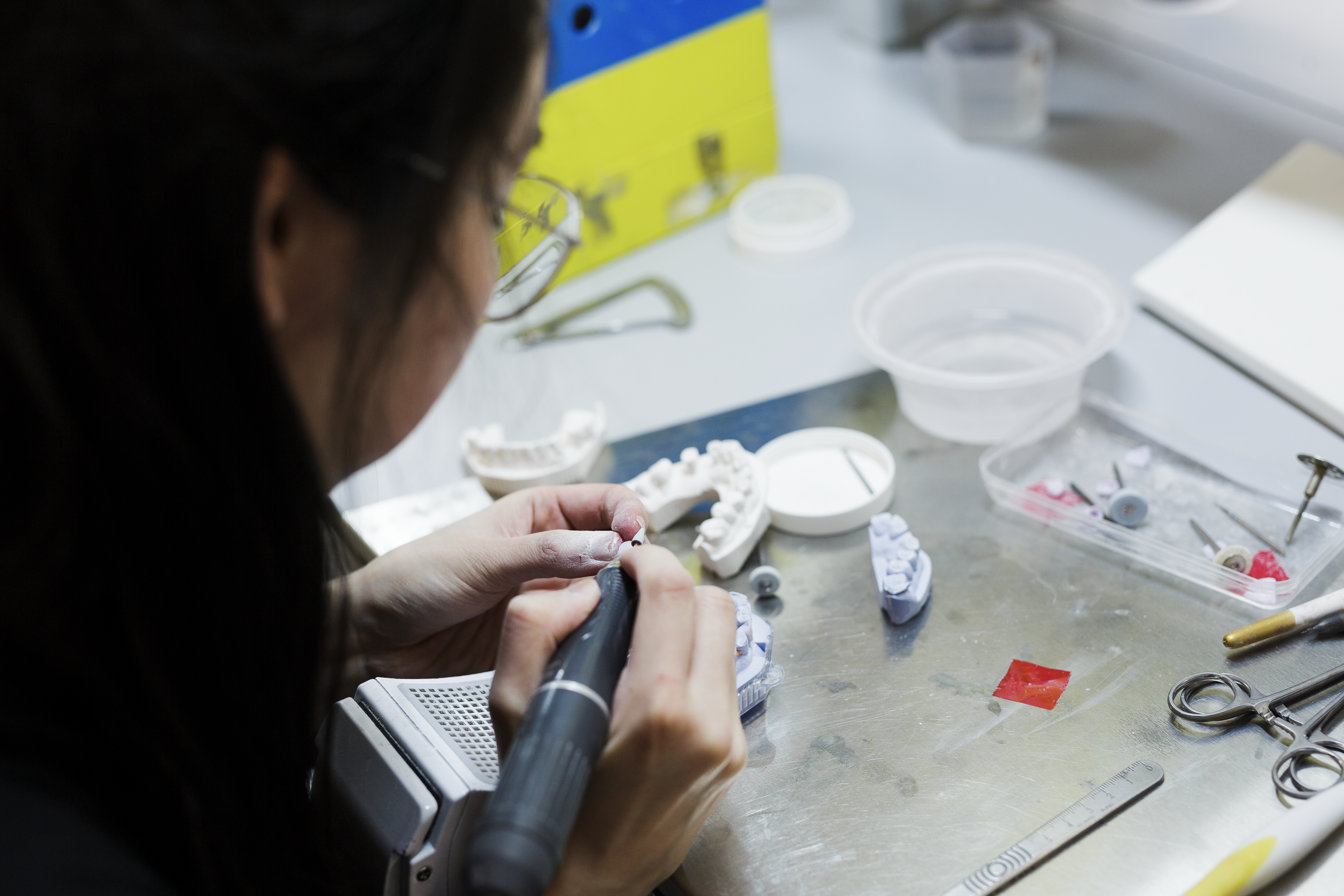 A dental laboratory technician uses a handpiece to work on dental prosthetics and models at a workstation, surrounded by various dental tools, materials, and equipment.