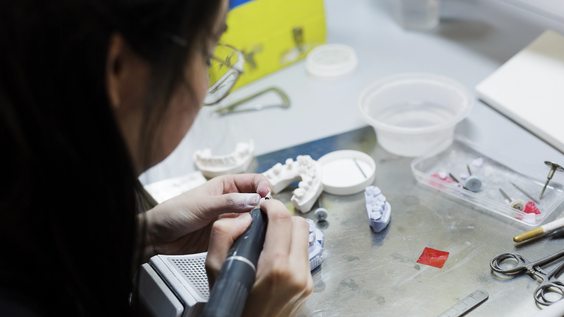 A dental laboratory technician uses a handpiece to work on dental prosthetics and models at a workstation, surrounded by various dental tools, materials, and equipment.