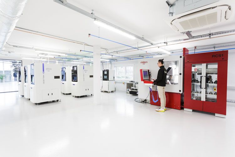 A technician operates advanced manufacturing equipment in a bright, clean laboratory space with white machines on the left and a prominent red ACSI machine on the right, featuring white fluorescent ceiling lights and blue electrical conduits.