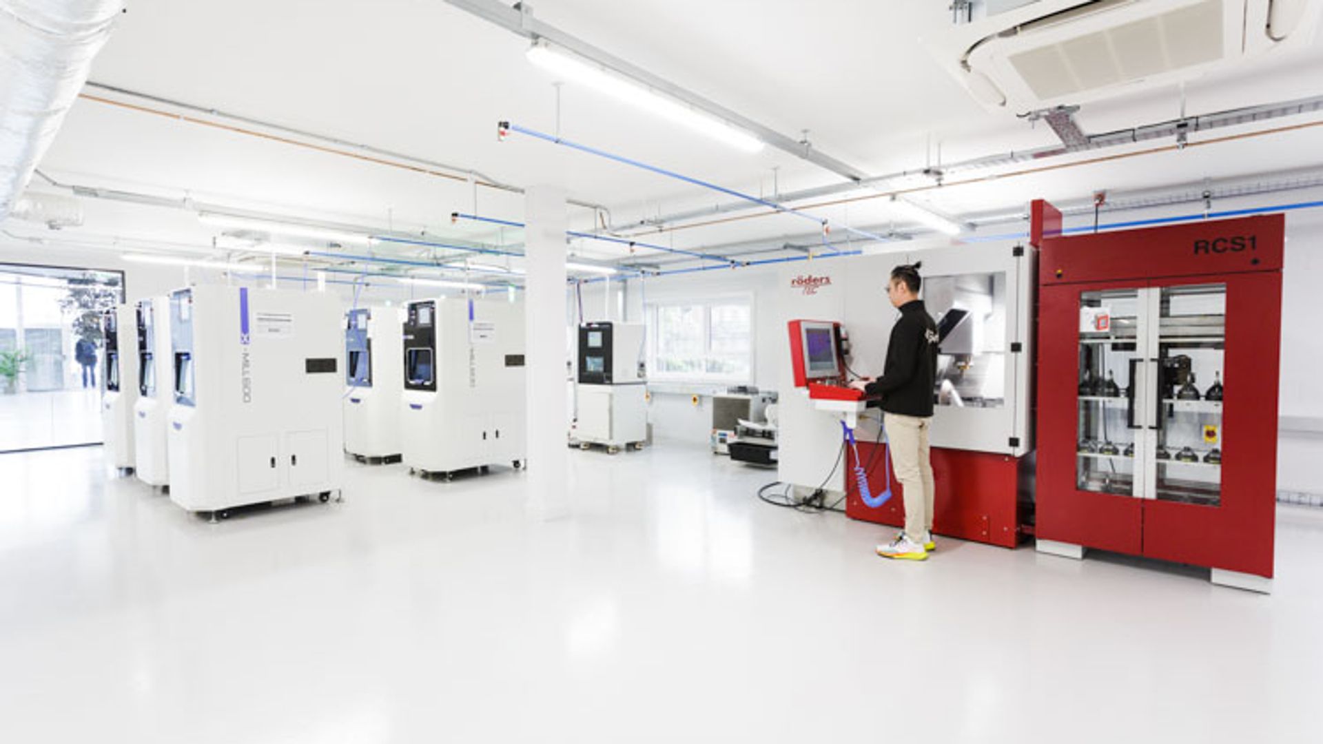 A technician operates advanced manufacturing equipment in a bright, clean laboratory space with white machines on the left and a prominent red ACSI machine on the right, featuring white fluorescent ceiling lights and blue electrical conduits.