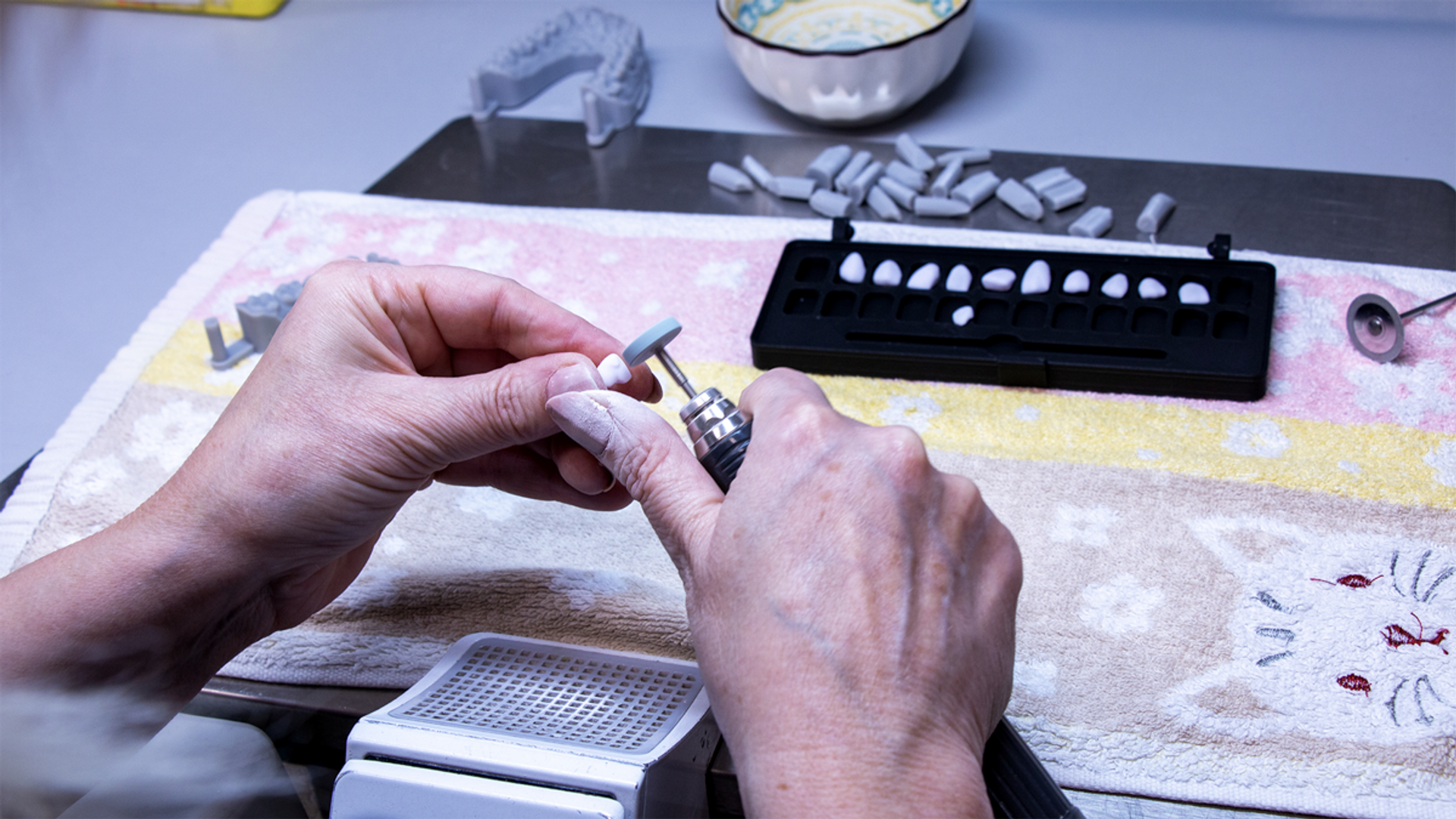 A dental technician's hands manipulate a shade guide while working with artificial teeth samples in a black tray, dental models, and laboratory materials on a work surface.