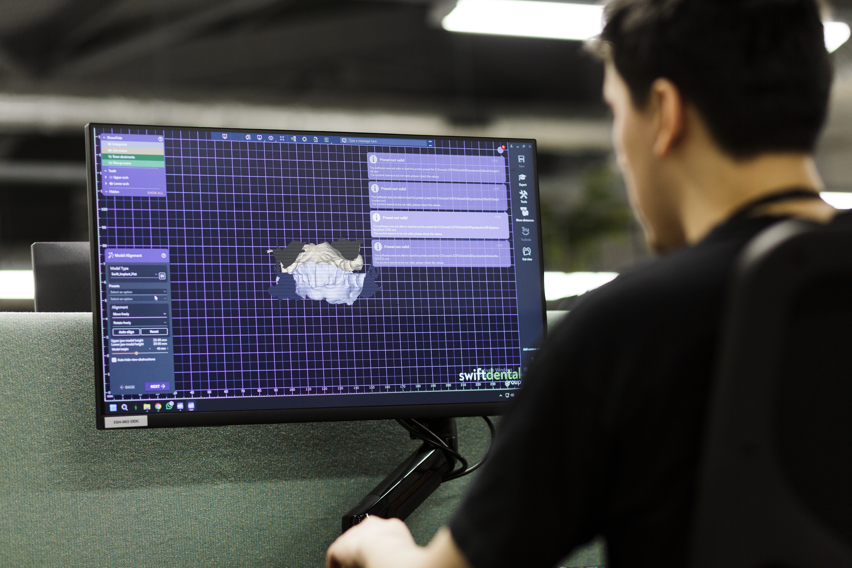A dental technician views a computer screen displaying Swift Dental software with a 3D digital model of a dental arch rendered on a blue grid interface for designing dental prosthetics and restorations.