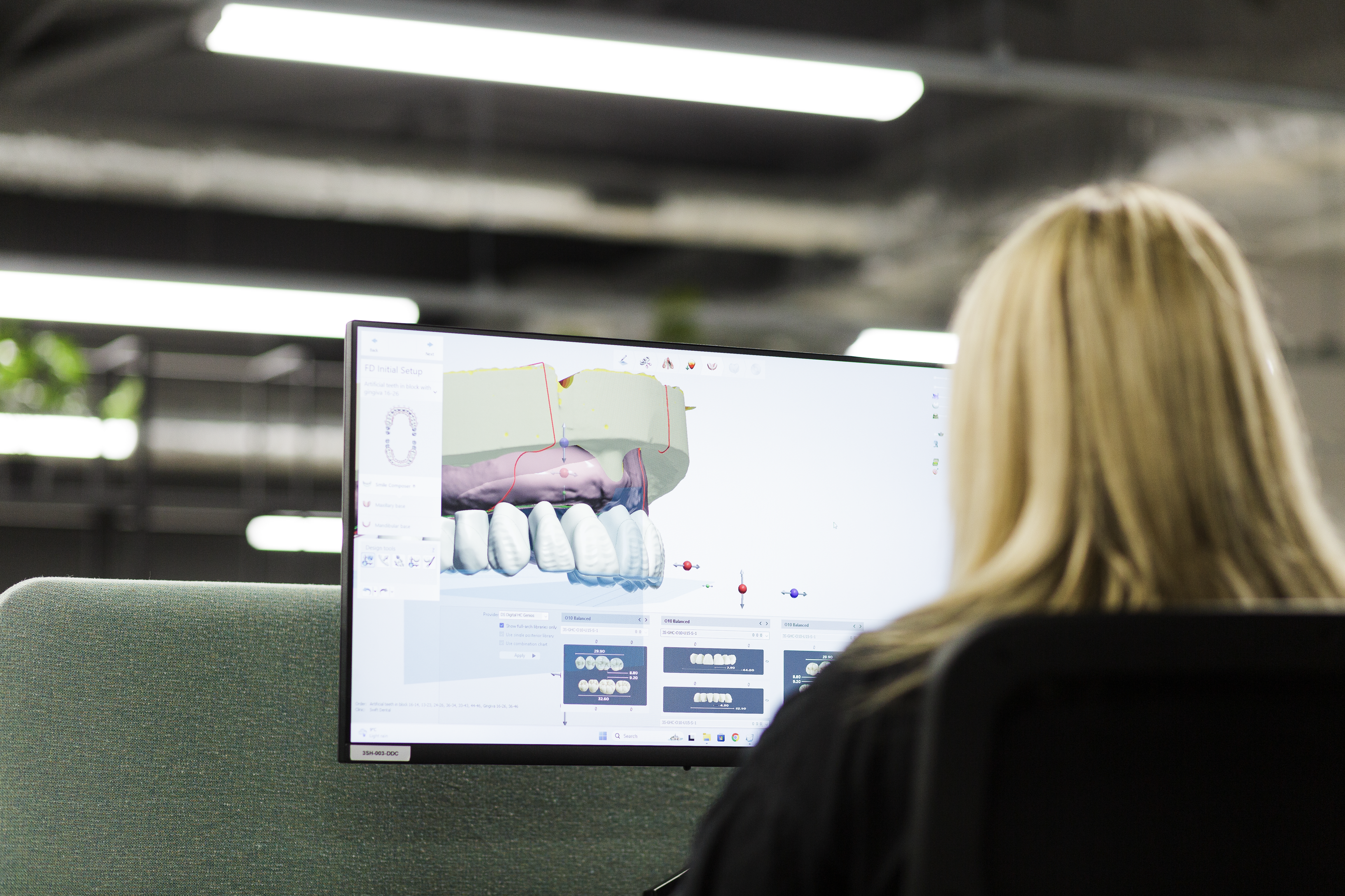 A dental technician views a computer monitor displaying 3D dental CAD software with a digital implant restoration design showing tooth crowns, bone structure, and bite plane analysis.