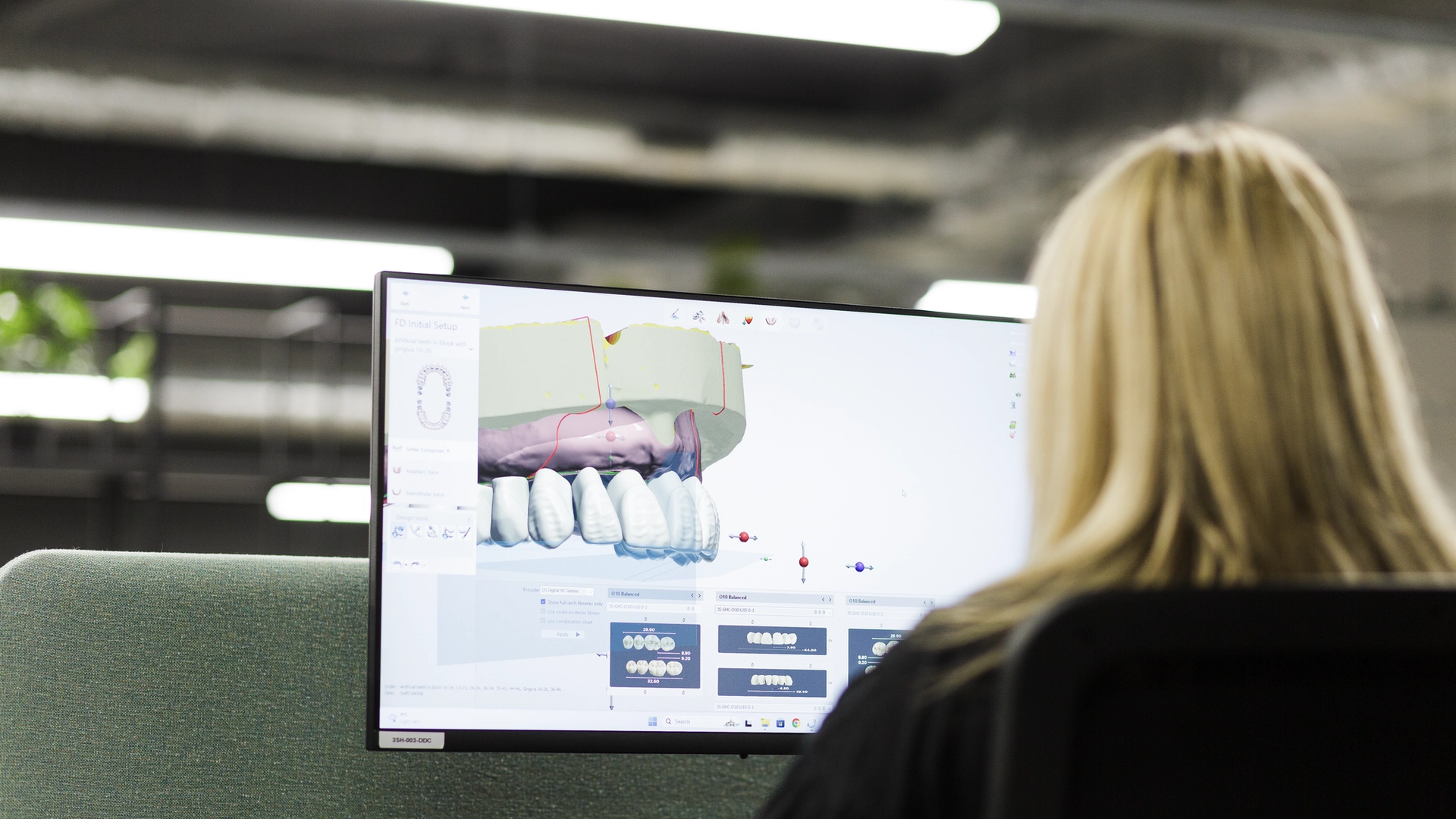 A dental technician views a computer monitor displaying 3D dental CAD software with a digital implant restoration design showing tooth crowns, bone structure, and bite plane analysis.