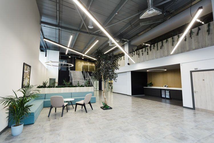 A spacious contemporary office lobby featuring a blue seating area, polished marble flooring, industrial ceiling with linear LED lighting, living plant installations, a reception desk, and a two-story interior with metal stairs and greenery.