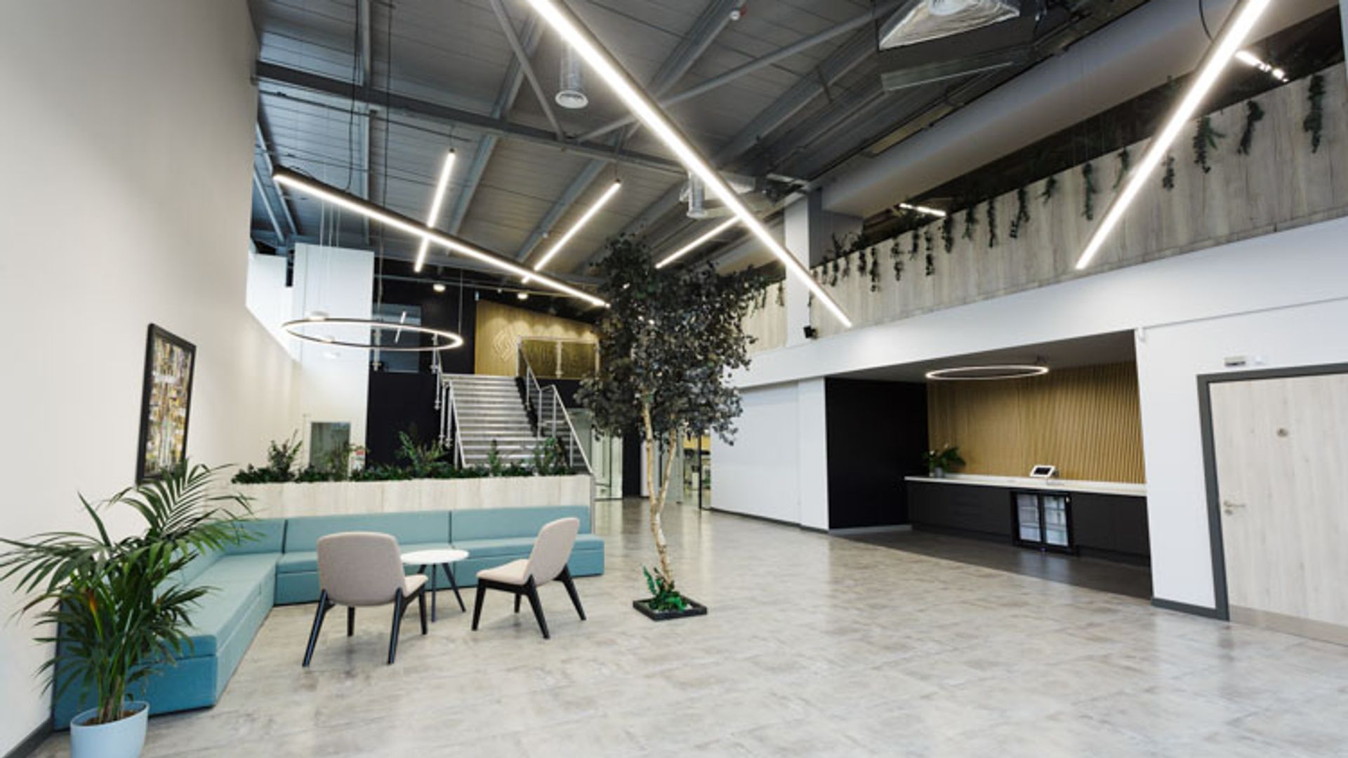 A spacious contemporary office lobby featuring a blue seating area, polished marble flooring, industrial ceiling with linear LED lighting, living plant installations, a reception desk, and a two-story interior with metal stairs and greenery.