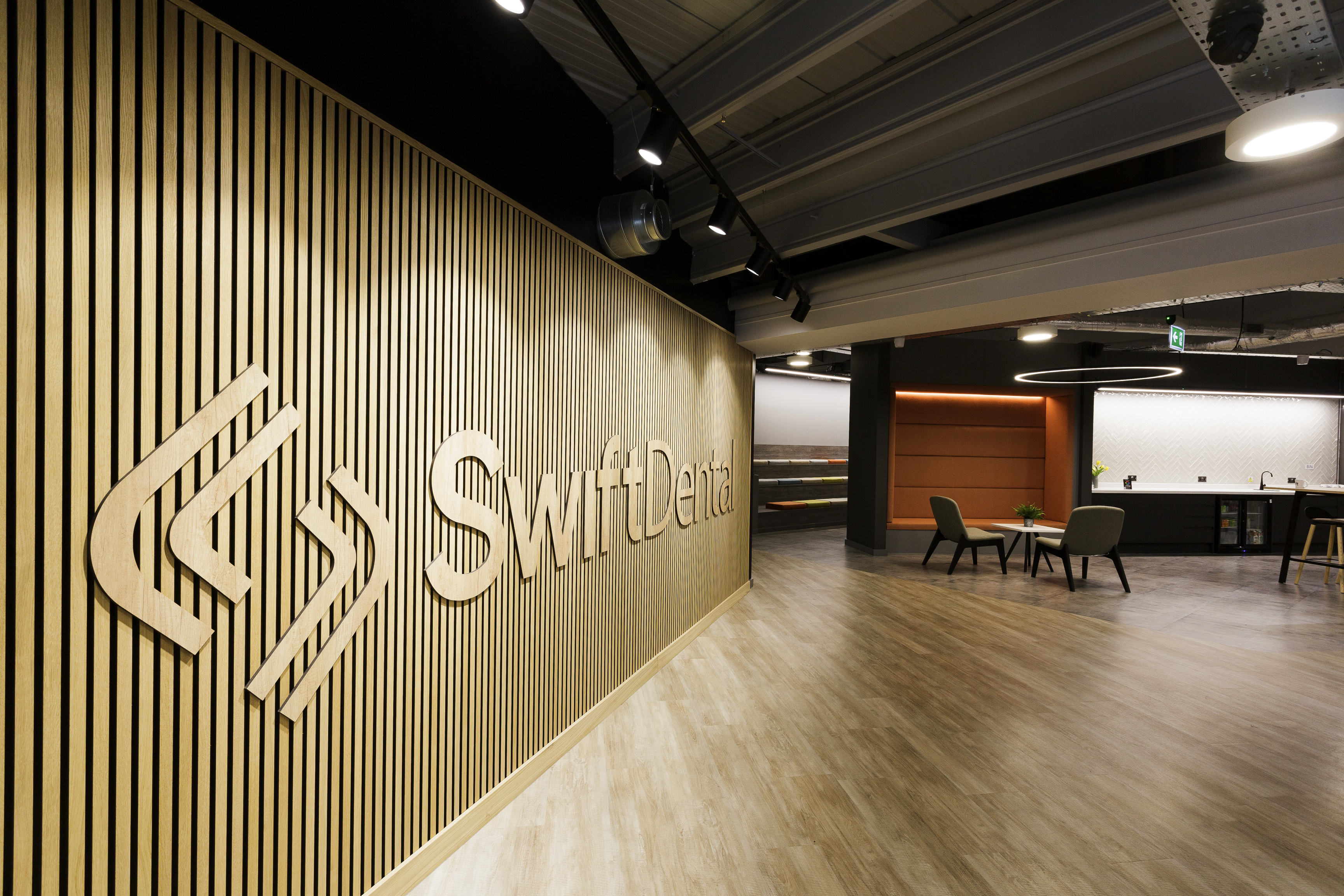 A contemporary dental practice entrance featuring a large brand logo on a textured wall panel, with a modern reception area, comfortable seating, and professional clinical spaces visible in the background.