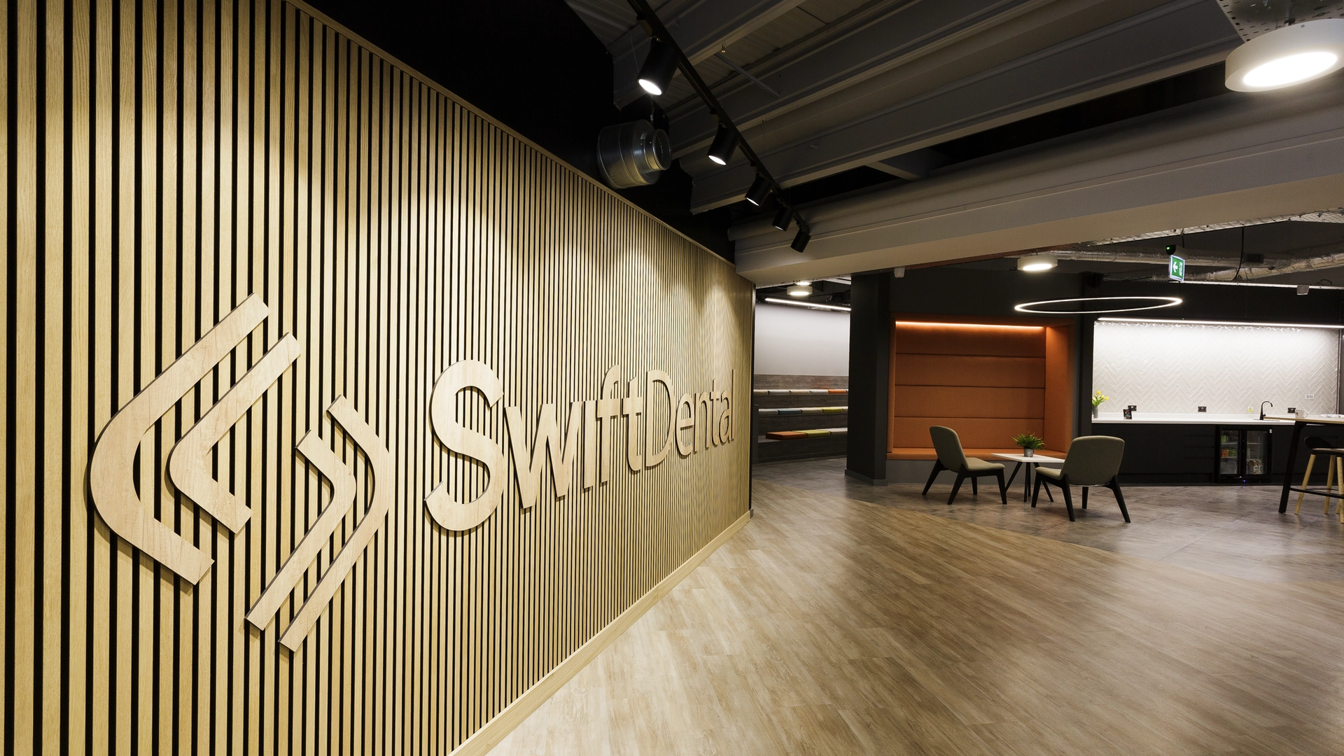 A contemporary dental practice entrance featuring a large brand logo on a textured wall panel, with a modern reception area, comfortable seating, and professional clinical spaces visible in the background.