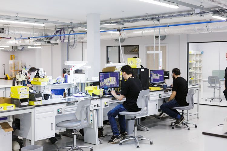 A bright, modern laboratory facility with multiple workstations where technicians in black shirts work at computer terminals, with scientific equipment, white cabinetry, and blue overhead piping visible throughout the clean, organized space.