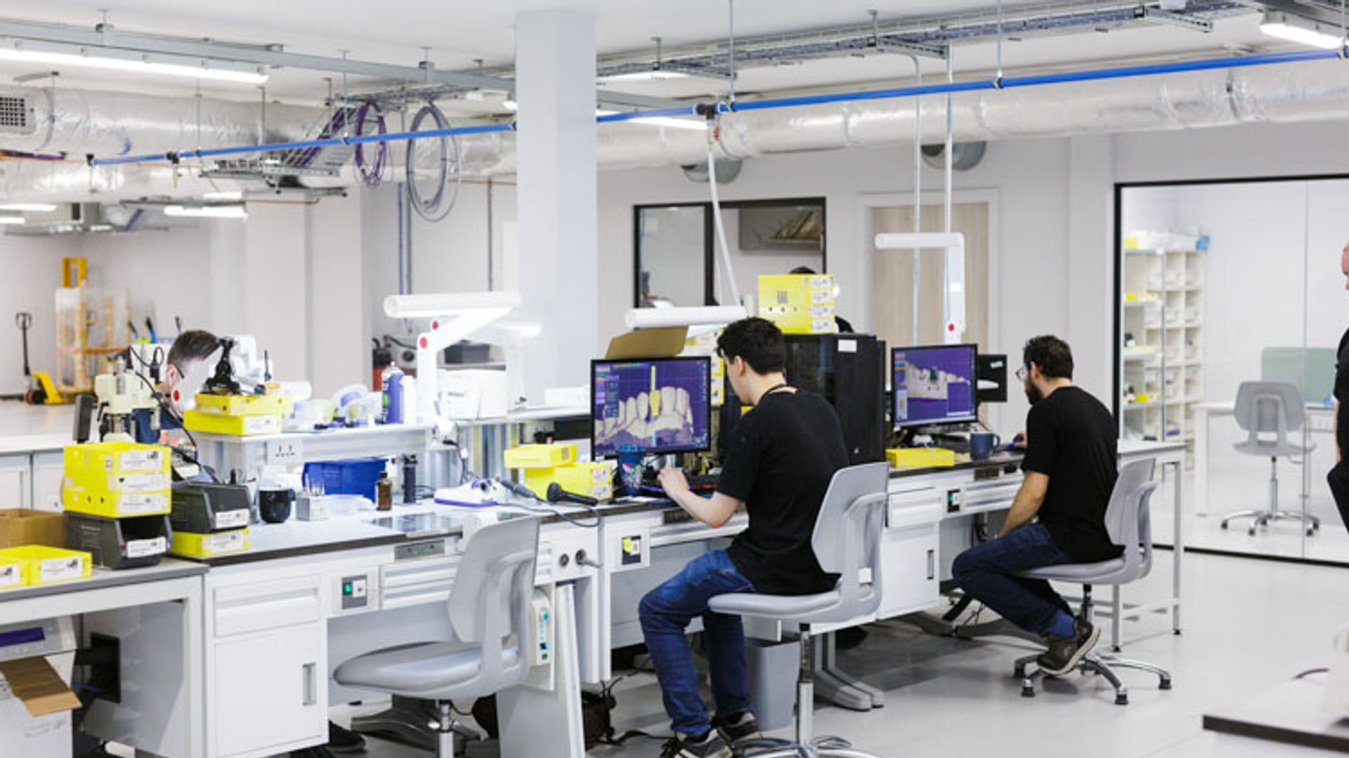 A bright, modern laboratory facility with multiple workstations where technicians in black shirts work at computer terminals, with scientific equipment, white cabinetry, and blue overhead piping visible throughout the clean, organized space.