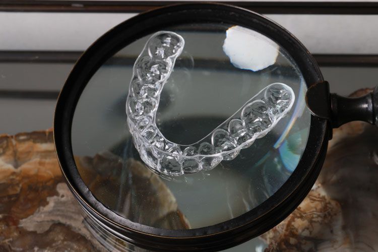 A clear plastic dental retainer is magnified under a large handheld magnifying glass, displaying the detailed structure of the orthodontic appliance with a small piece of white material visible in the lens.