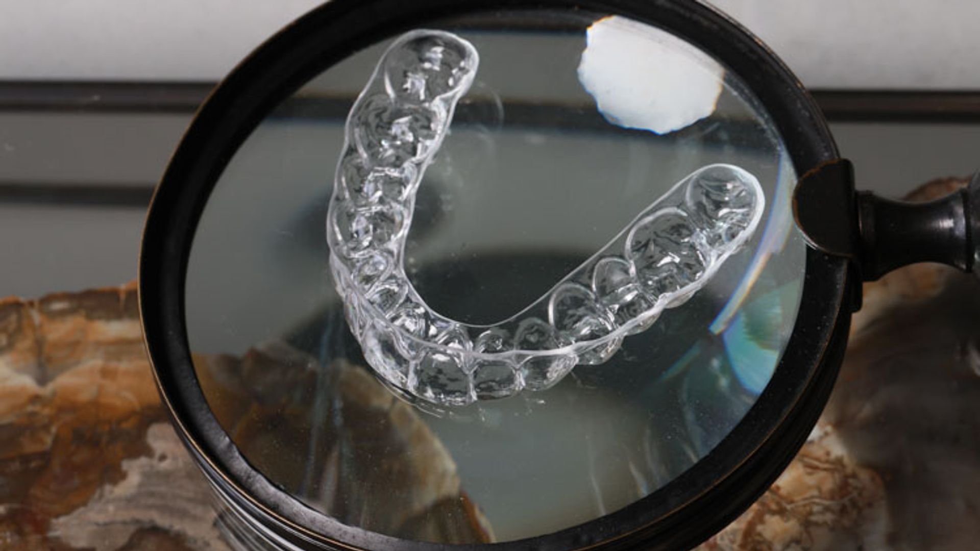 A clear plastic dental retainer is magnified under a large handheld magnifying glass, displaying the detailed structure of the orthodontic appliance with a small piece of white material visible in the lens.