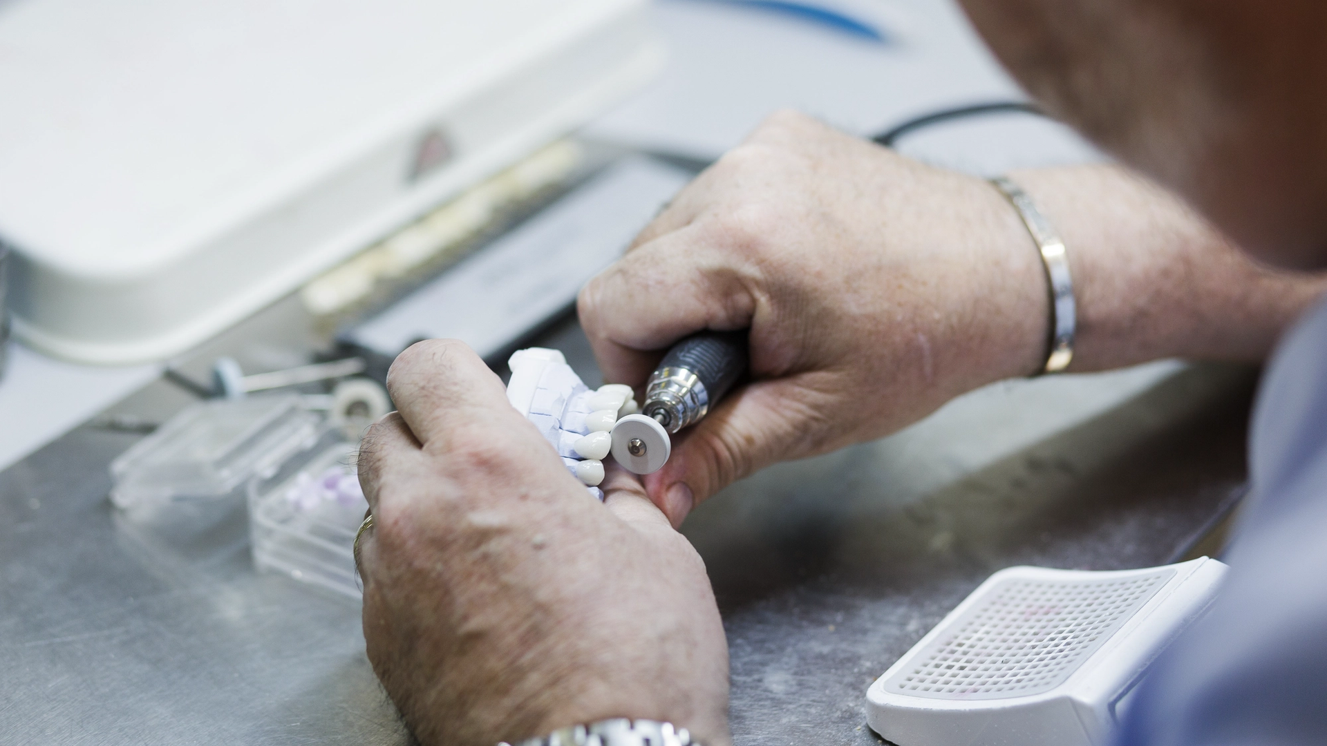 A dental technician holds a white ceramic zirconia abutment attached to a dental implant handpiece during laboratory fabrication of a tooth restoration.