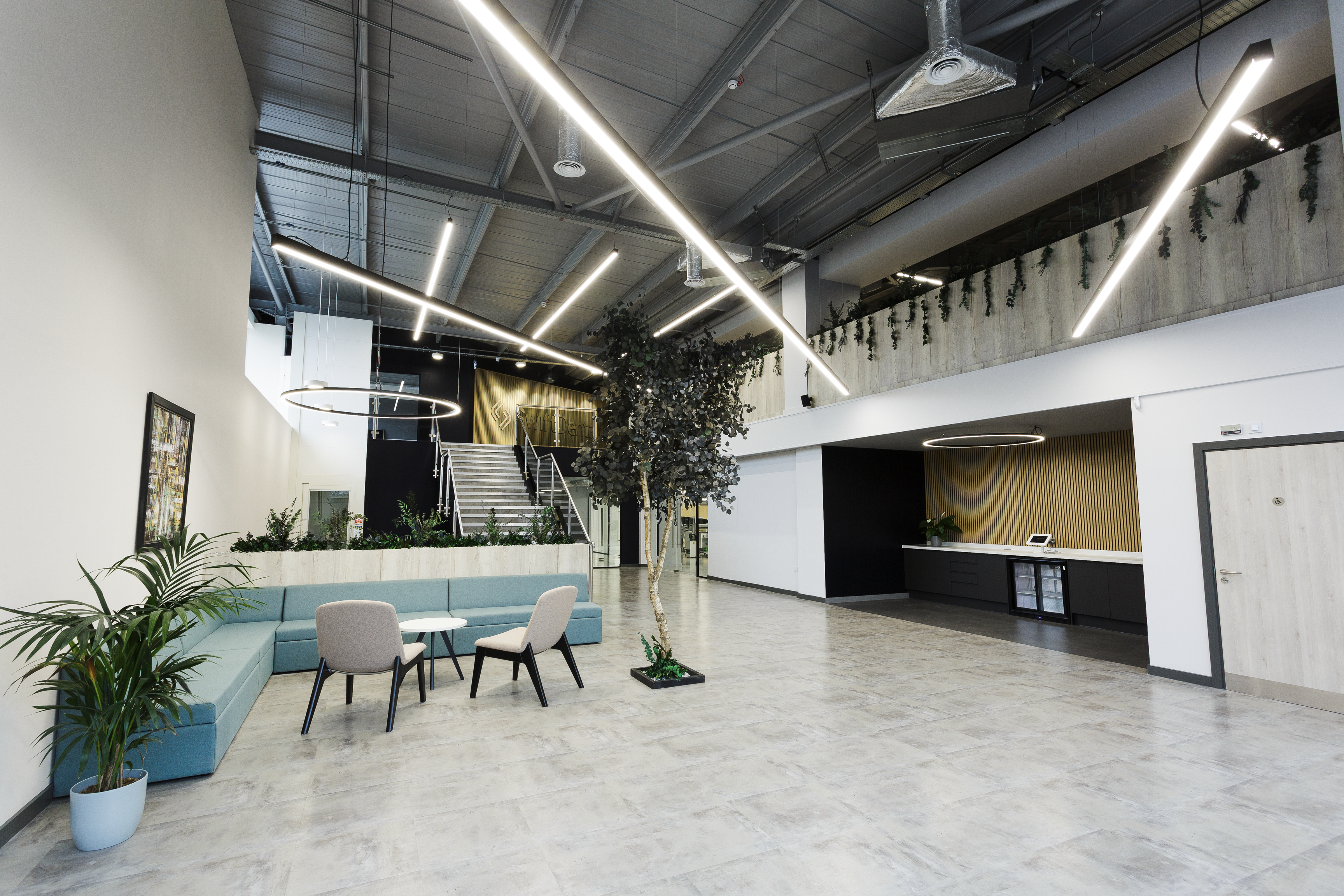 A spacious, contemporary dental laboratory reception area featuring polished concrete flooring, bright linear ceiling lights, a blue modular seating area, and a black reception desk with gold accents.