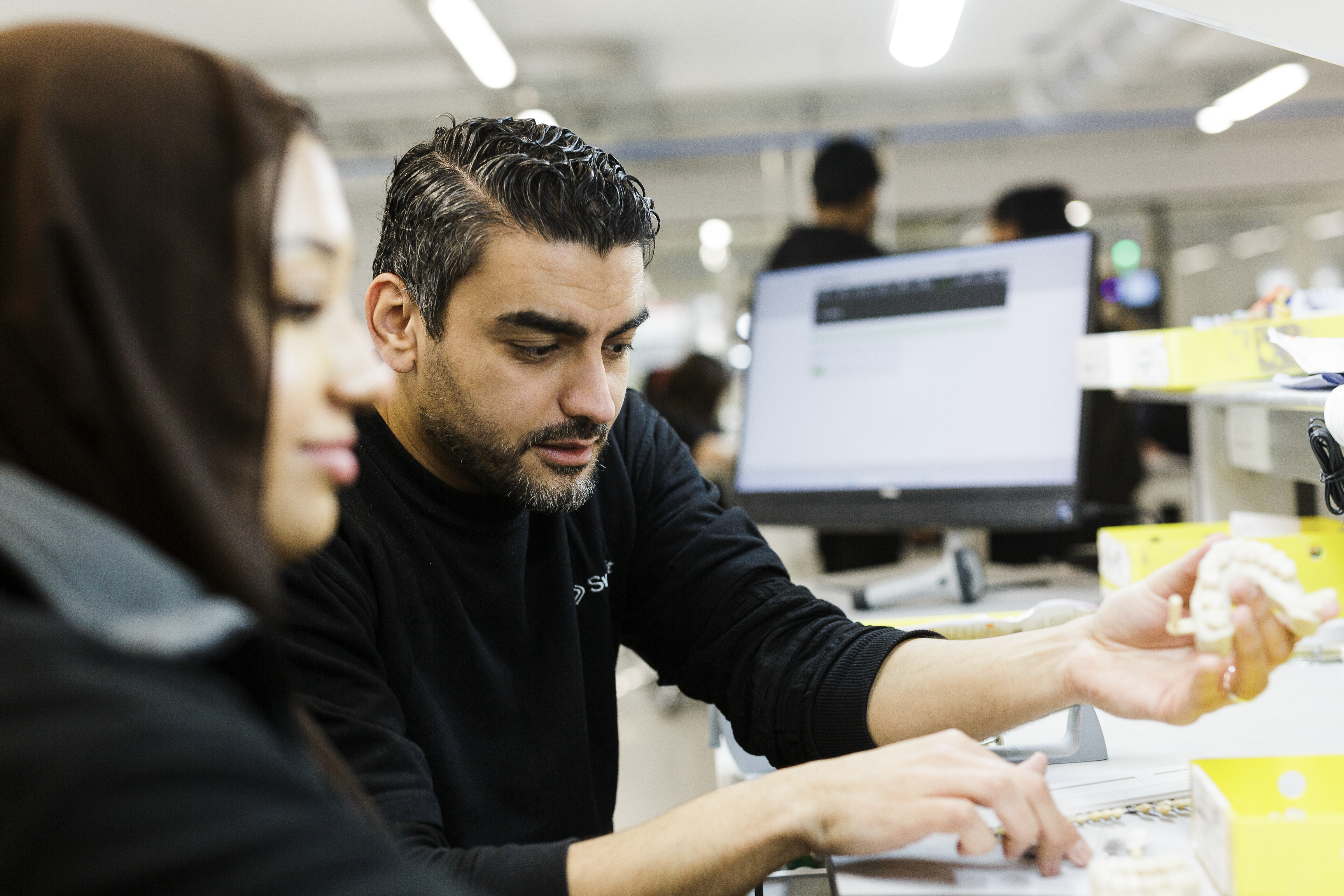 A dental technician in a laboratory setting examines a dental prosthetic model while consulting with a colleague, with computer workstations and organized workspace visible in the background.