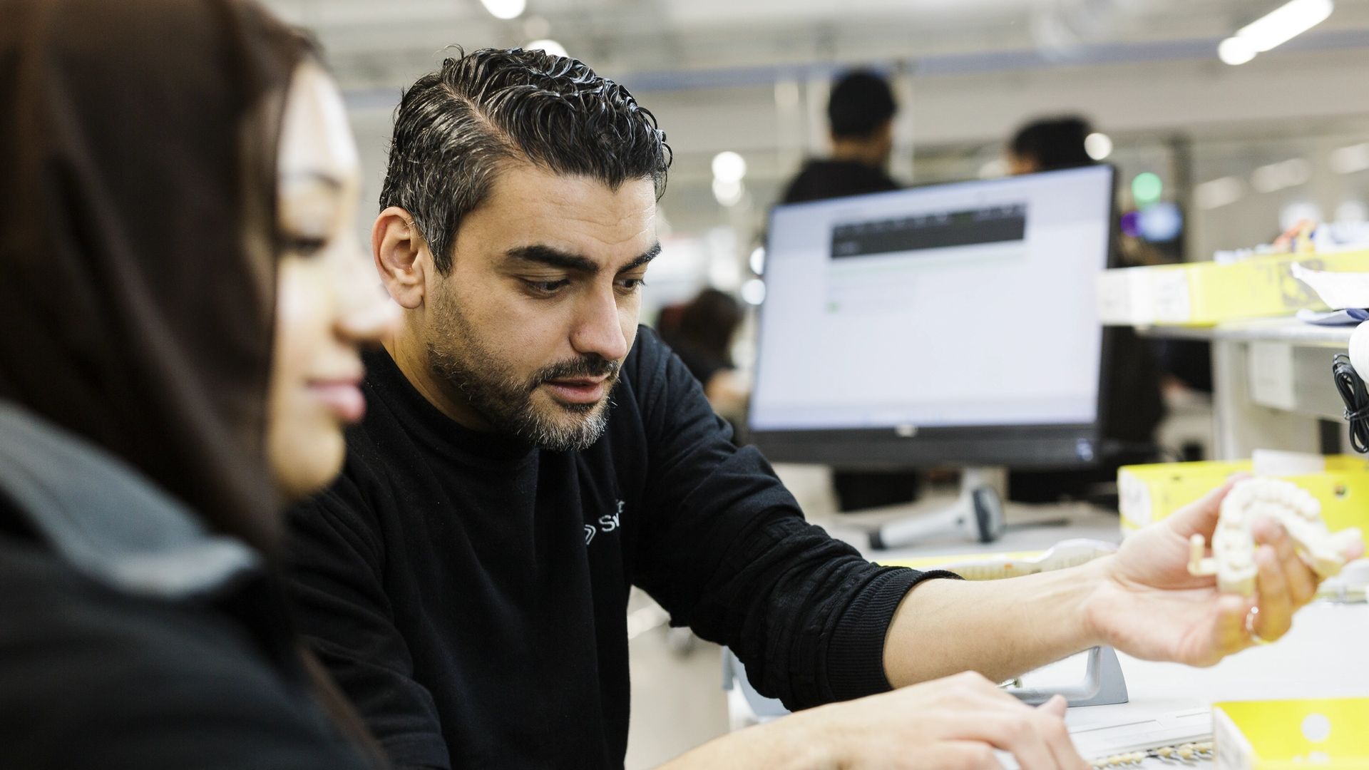 A dental technician in a laboratory setting examines a dental prosthetic model while consulting with a colleague, with computer workstations and organized workspace visible in the background.