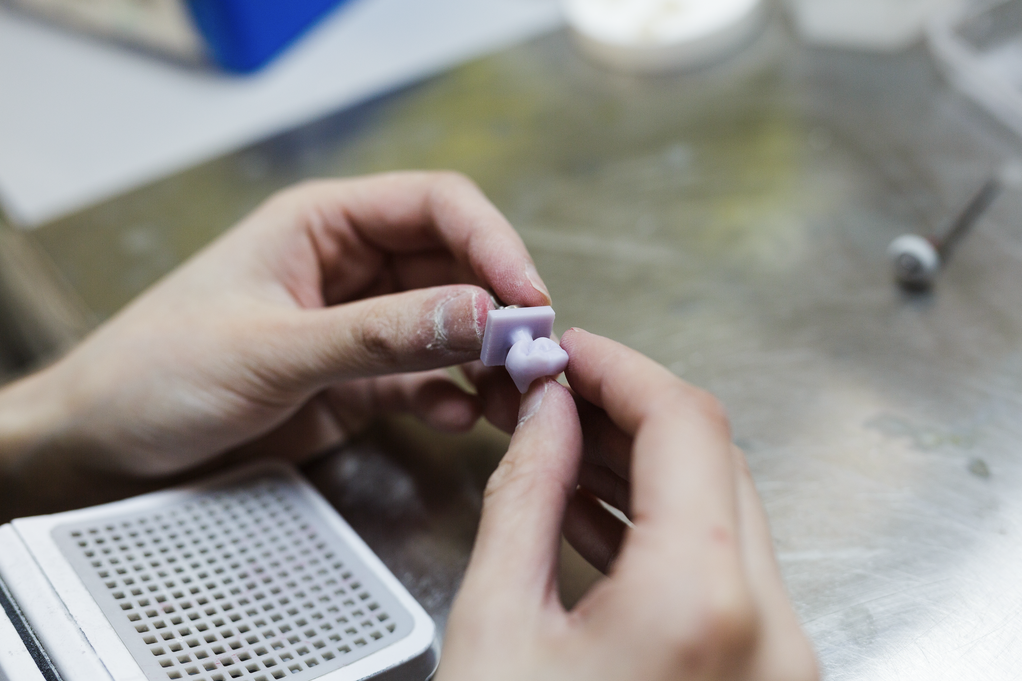 A dental technician holds a small light-colored composite resin sample between their fingers for examination, with a perforated shade guide tray visible in the lower left corner of the laboratory workspace.
