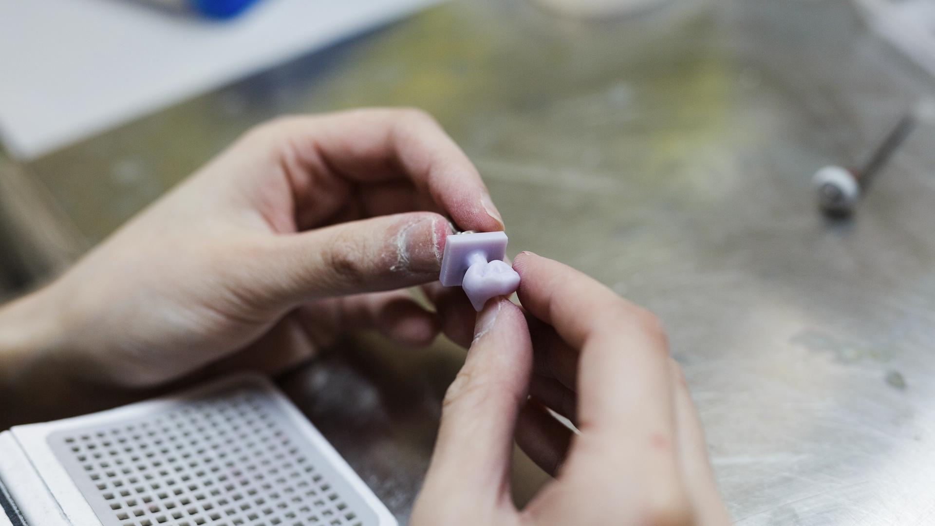 A dental technician holds a small light-colored composite resin sample between their fingers for examination, with a perforated shade guide tray visible in the lower left corner of the laboratory workspace.