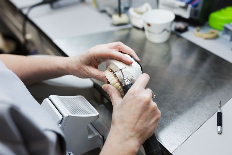 A dental technician examines a white dental model or prosthetic device with their hands while working at a laboratory bench with various tools and materials visible in the background.
