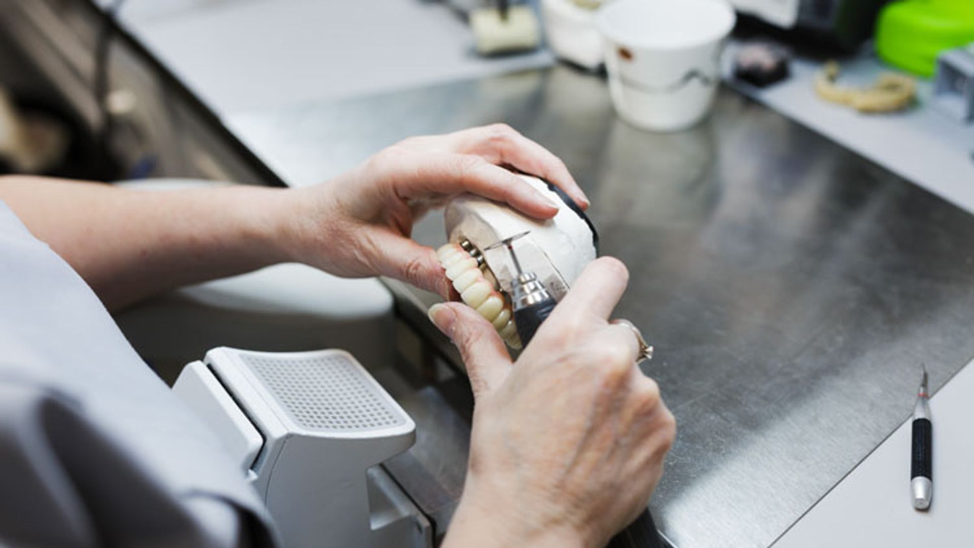 A dental technician examines a white dental model or prosthetic device with their hands while working at a laboratory bench with various tools and materials visible in the background.