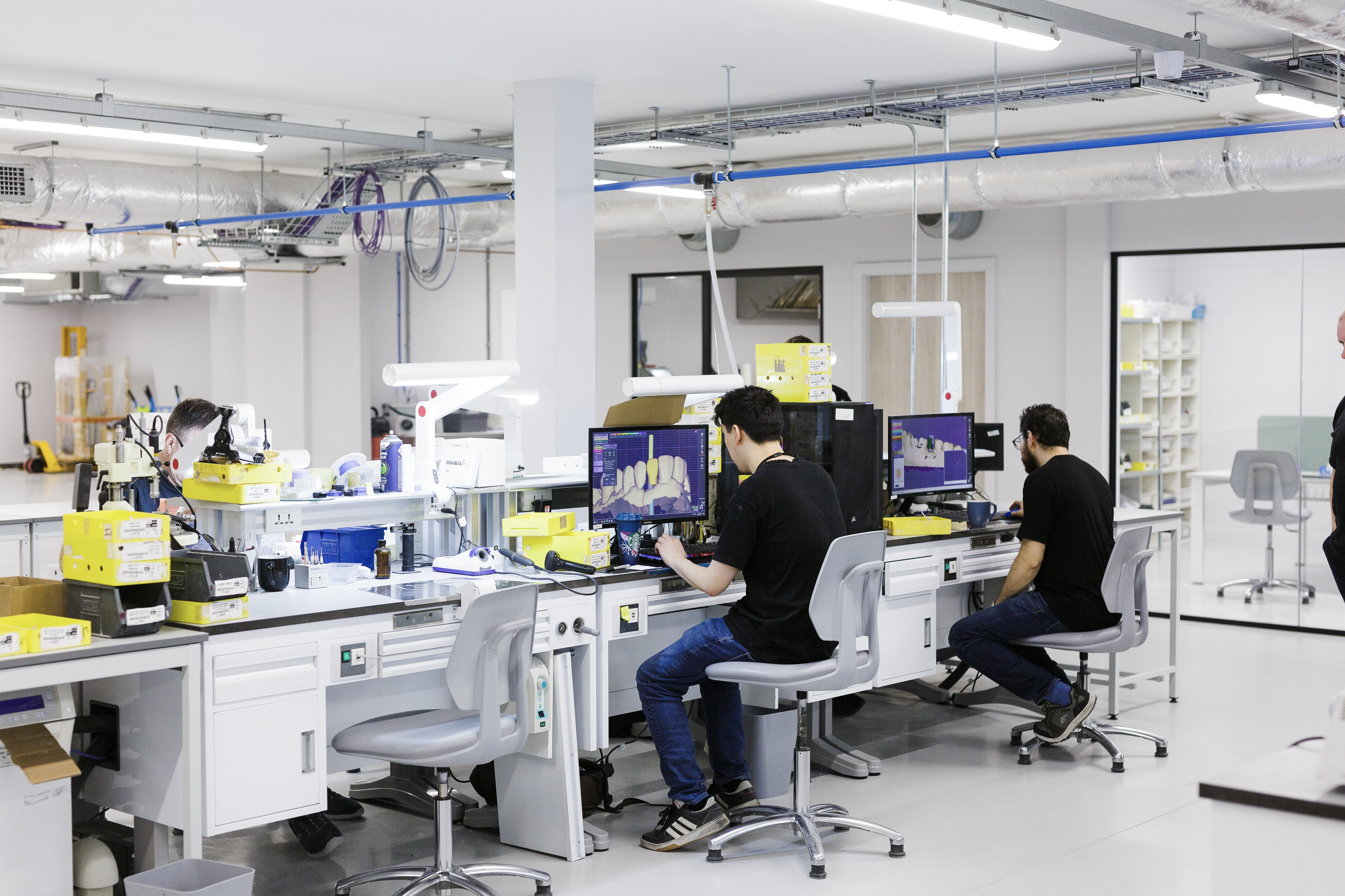Dental technicians work at computer workstations in a modern laboratory, viewing digital 3D tooth designs on monitors while operating CAD/CAM milling equipment and digital scanning systems.