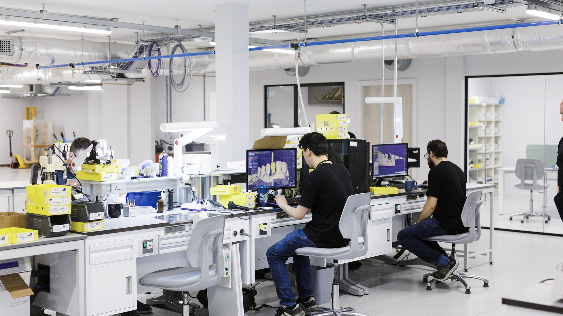 Dental technicians work at computer workstations in a modern laboratory, viewing digital 3D tooth designs on monitors while operating CAD/CAM milling equipment and digital scanning systems.