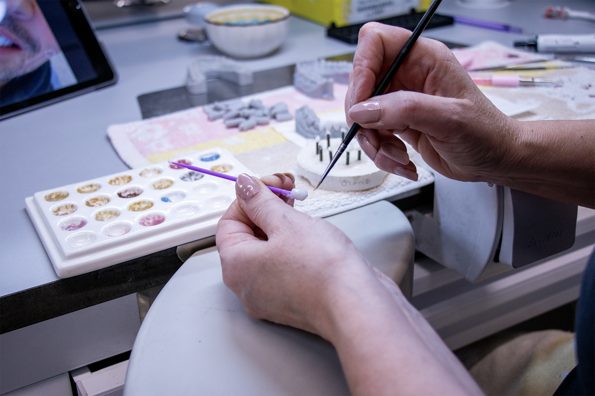 A dental technician uses a fine brush to apply tinted composite materials to a dental restoration model while referencing a comprehensive shade guide palette containing multiple tooth color samples.