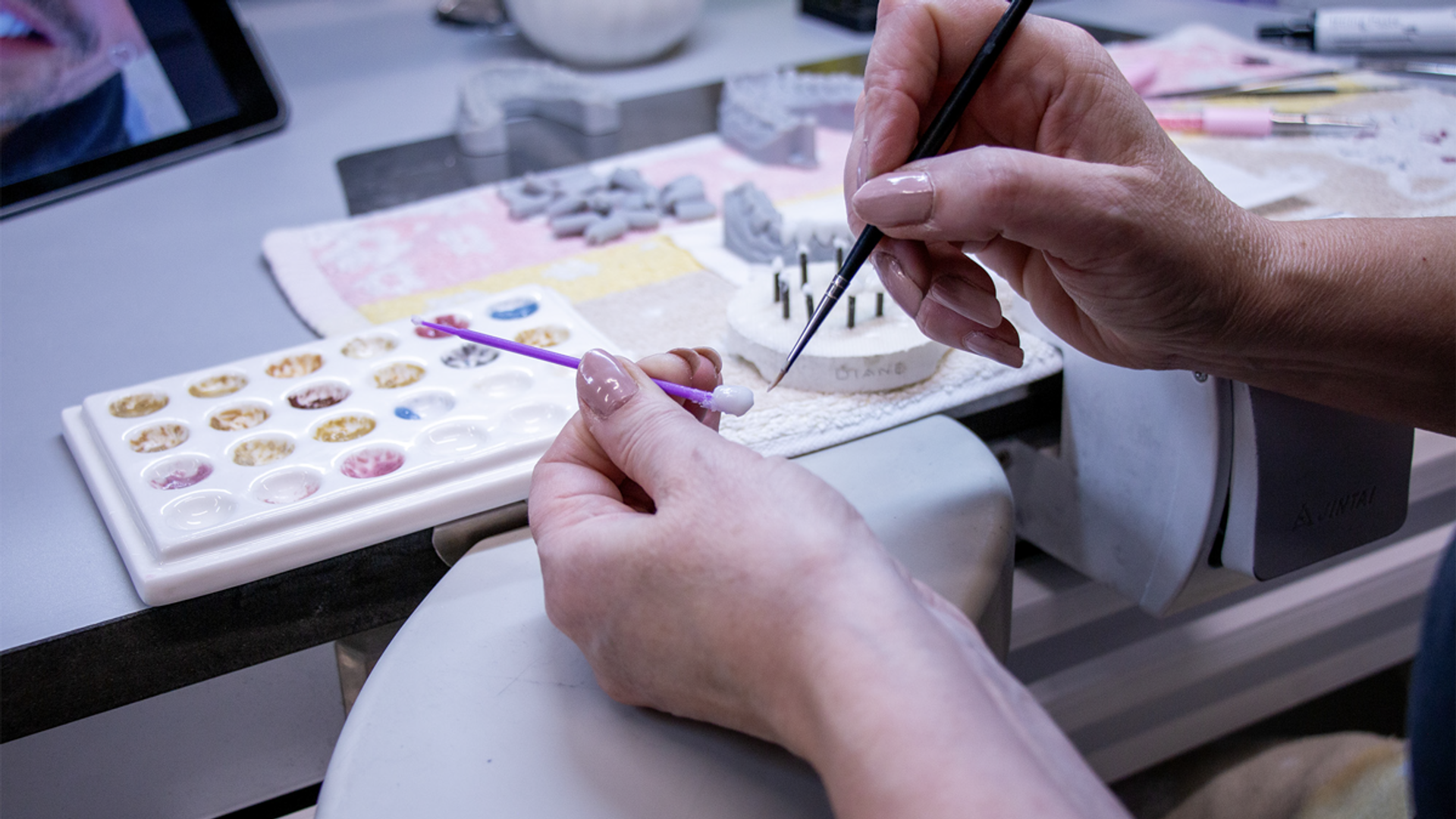 A dental technician uses a fine brush to apply tinted composite materials to a dental restoration model while referencing a comprehensive shade guide palette containing multiple tooth color samples.