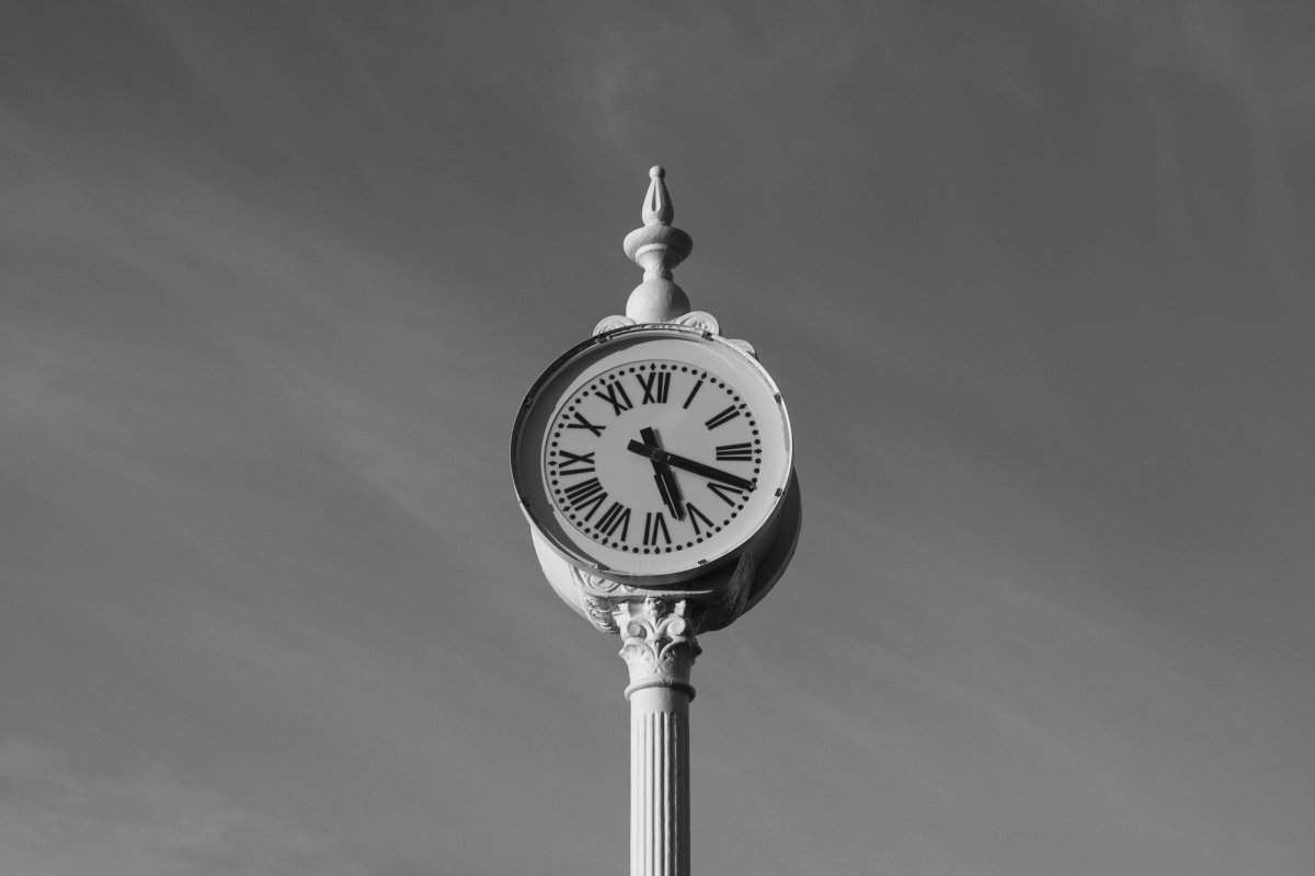 Black and white photograph of a vintage outdoor street clock mounted on an ornate pole against a clear sky, symbolizing time and punctuality.