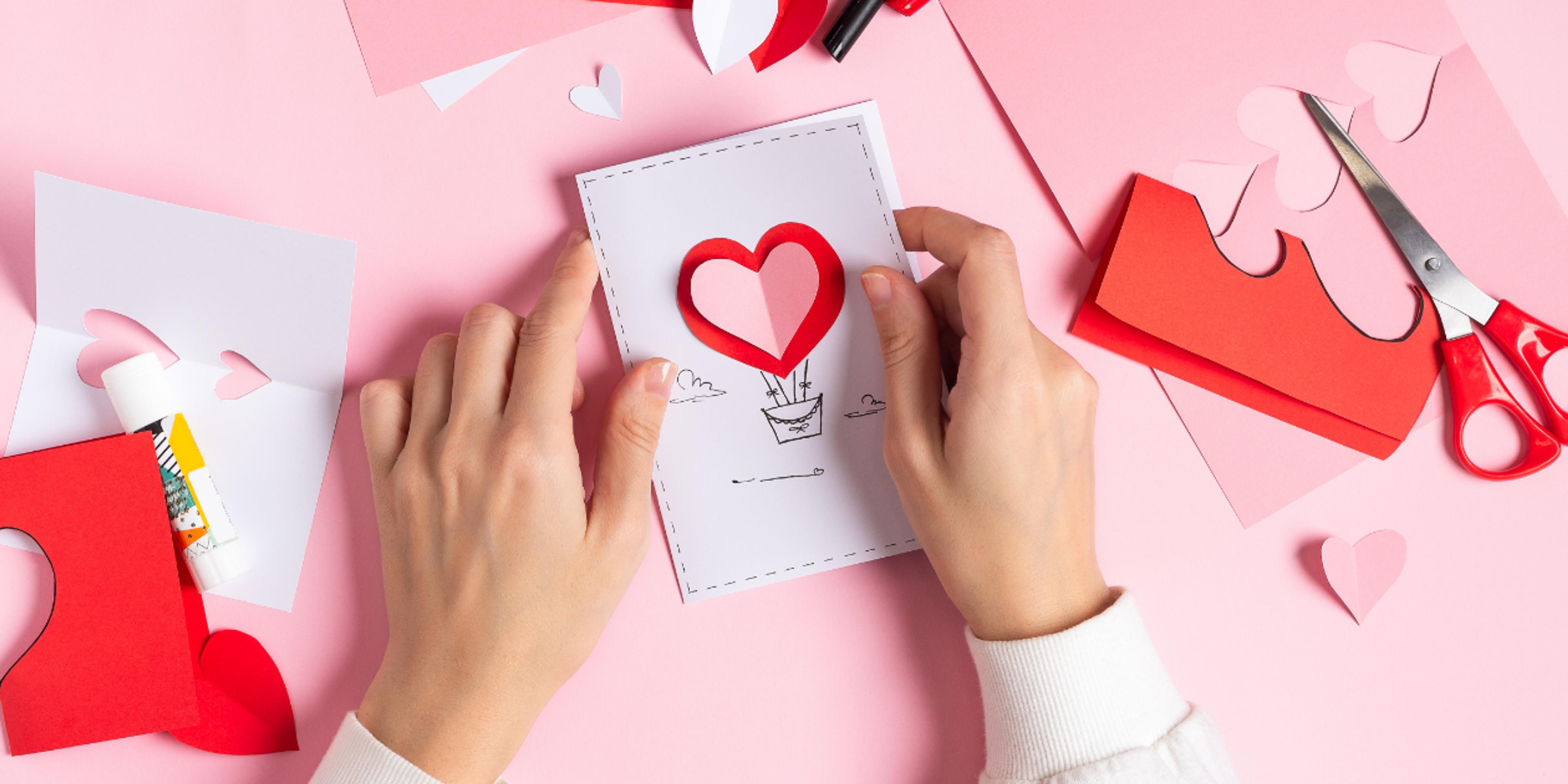 hands making a valentines day card on a pink background