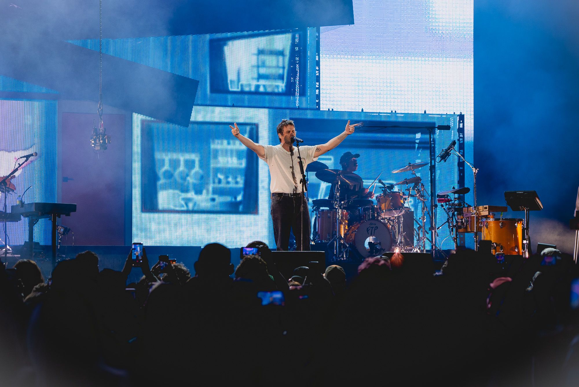 Mark Foster singing at a microphone stand with arms pointed outwards in a 'finger guns' gesture. Drummer can be seen behind him as well large LED wall showing graphic of house front.