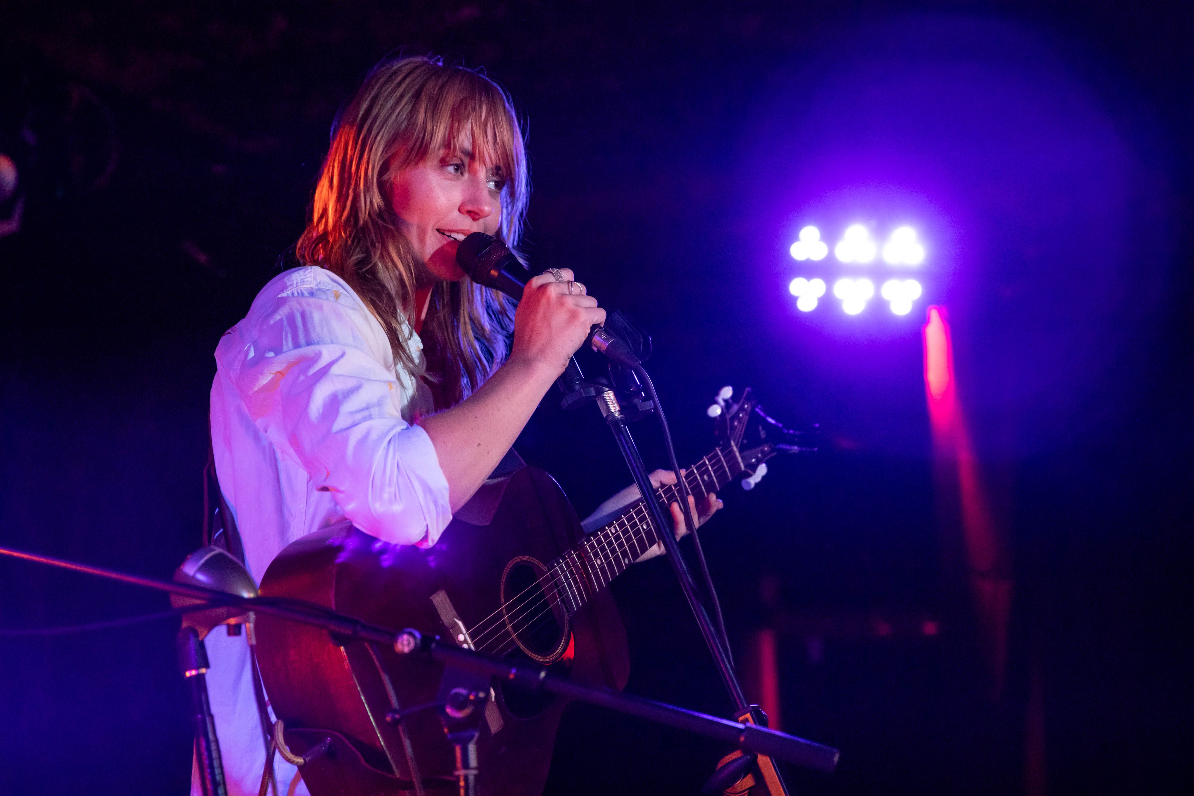 Gordi speaks into the microphone, bathed in purple light, with a guitar around her neck