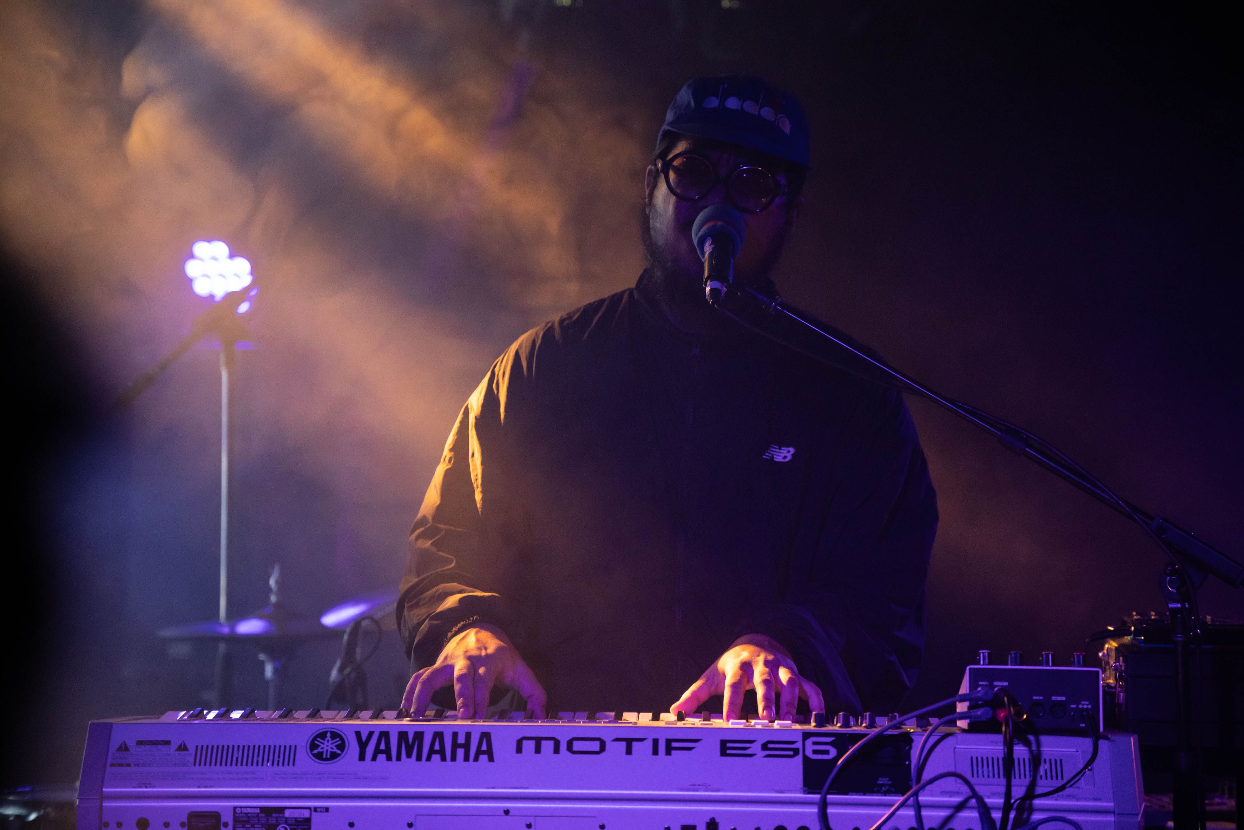 Mndsgn plays keyboard and sings under purple and yellow light