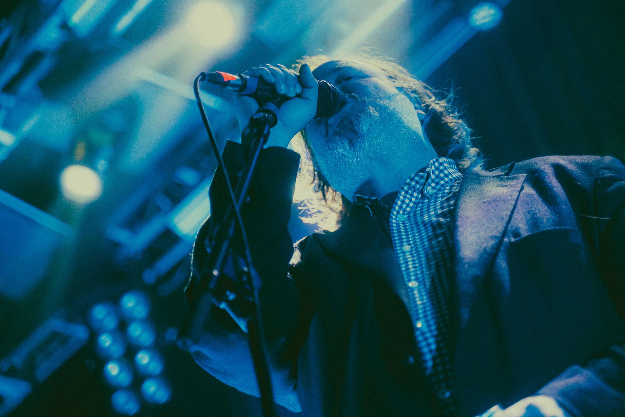 Protomartyr frontman Joe Casey is dressed in a button up shirt annd suit jacket, singing into a microphone in a low angle close up shot, washed in green and blue light.