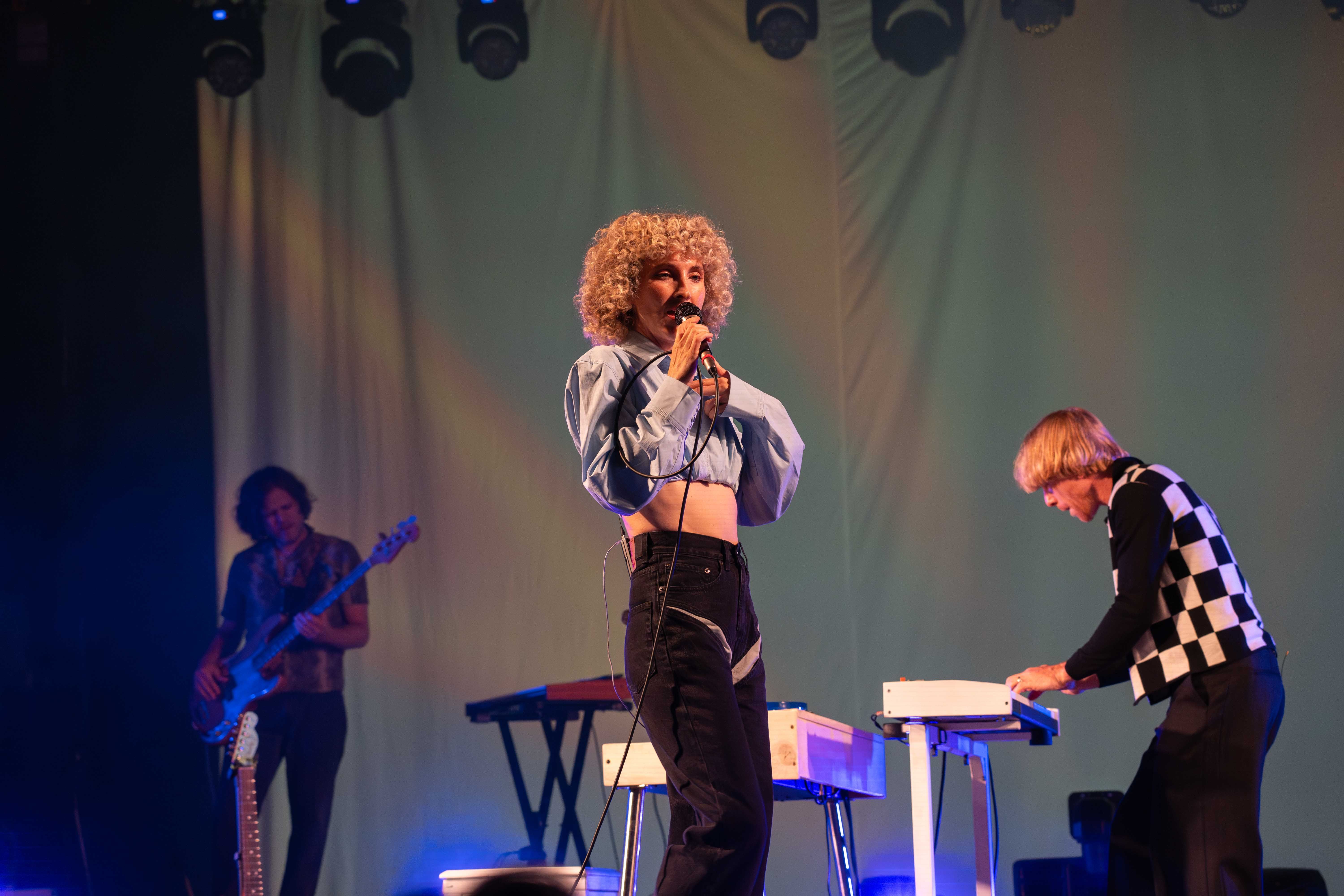 Alaina Moore of Tennis singing into a microphone on stage with Patrick Riley playing keyboard behind, and Ryan Tullock playing bass behind him