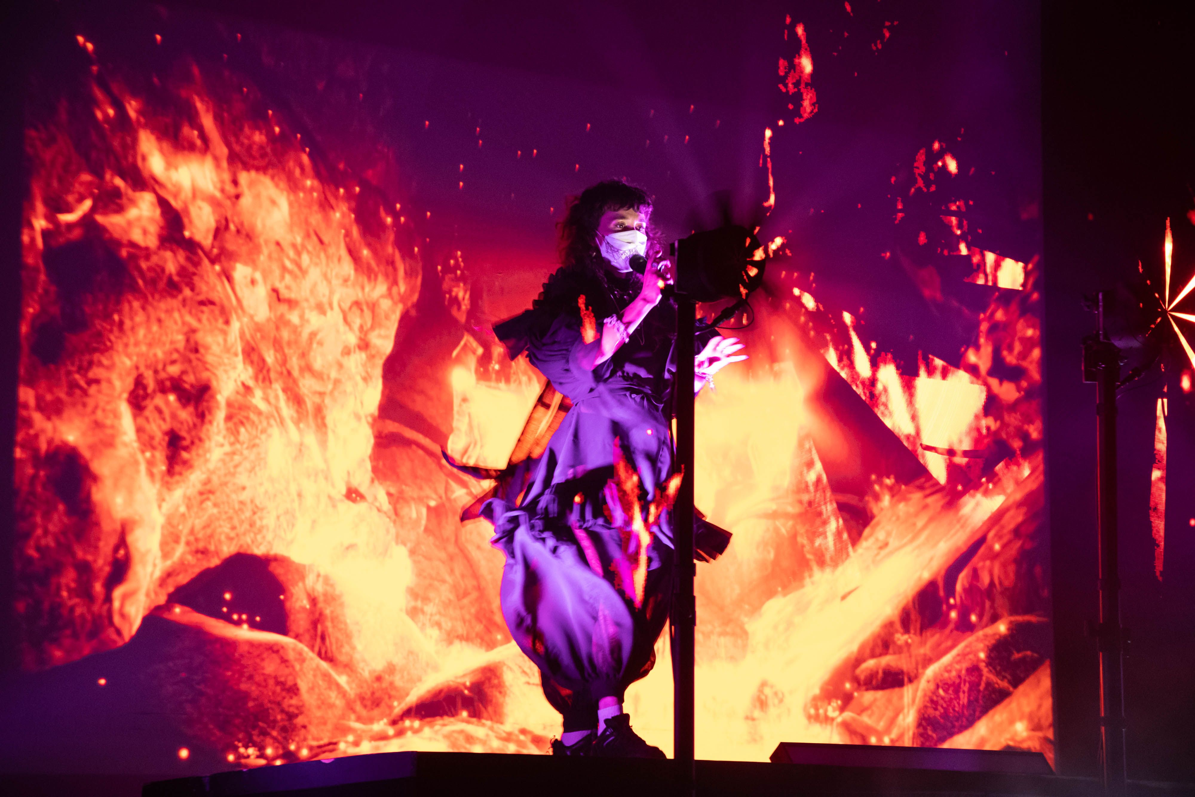 MJ from Purity Ring is surrounded by fire graphics behind and in front while singing into a wireless mic