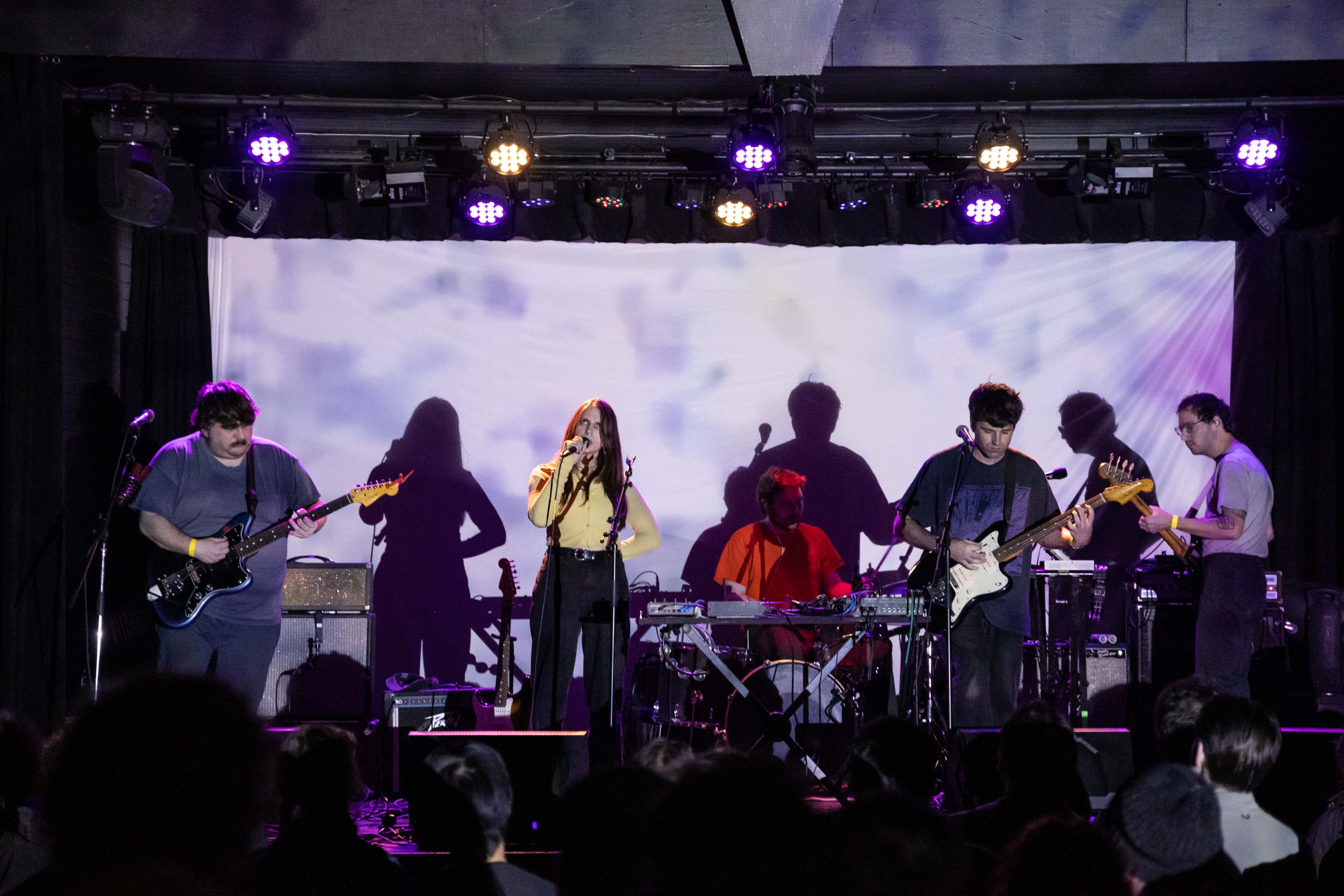 The full band of Dummy is on the stage at The Wise lit by a projection with a screen behind them
