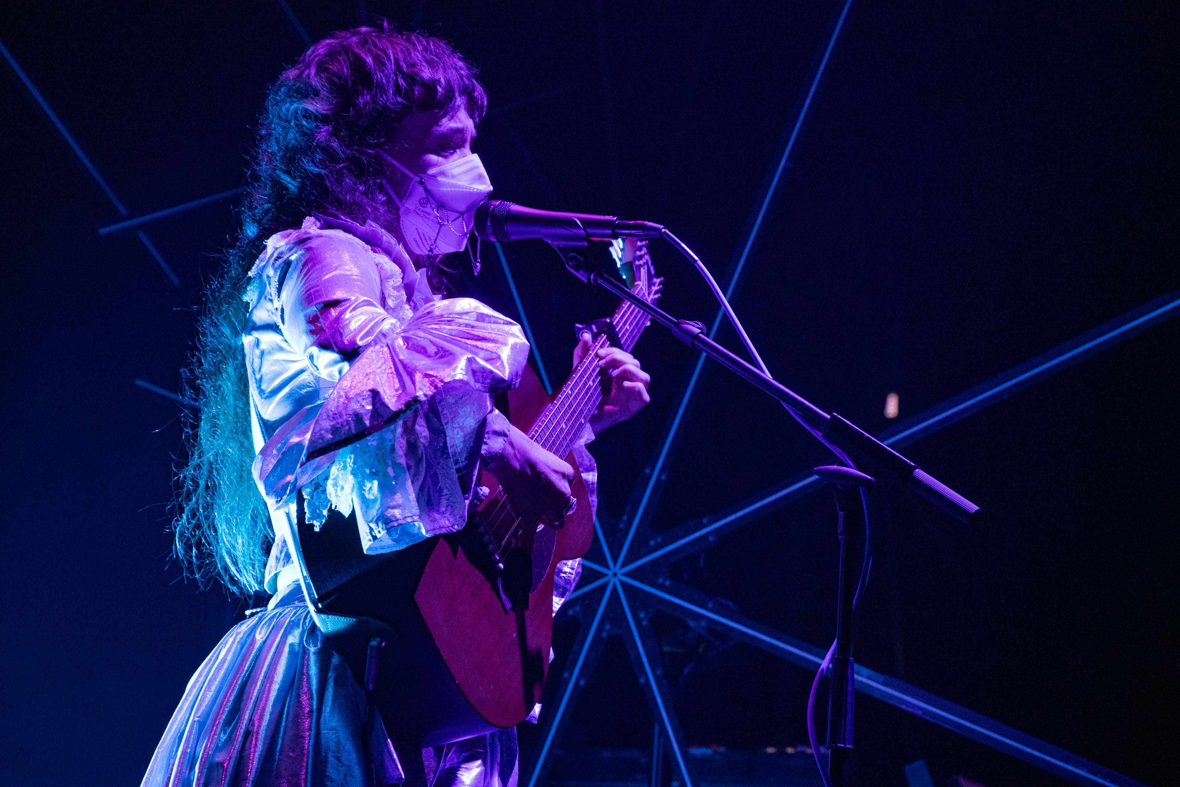 MMJ is lit in purple and blue, reflecting off her silver dress, holding an acoustic guitar