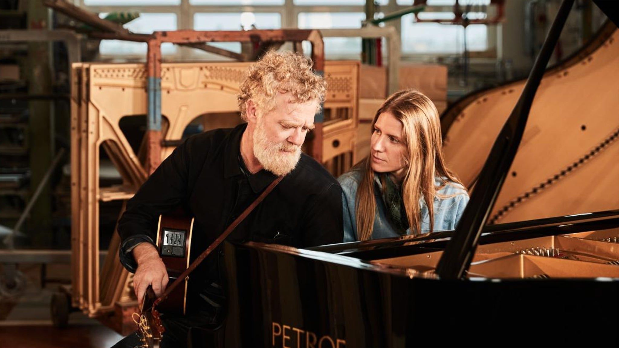 The Swell Season's Glen Hansard and Markéta Irglová sit together by a piano. Hansard holds a guitar.