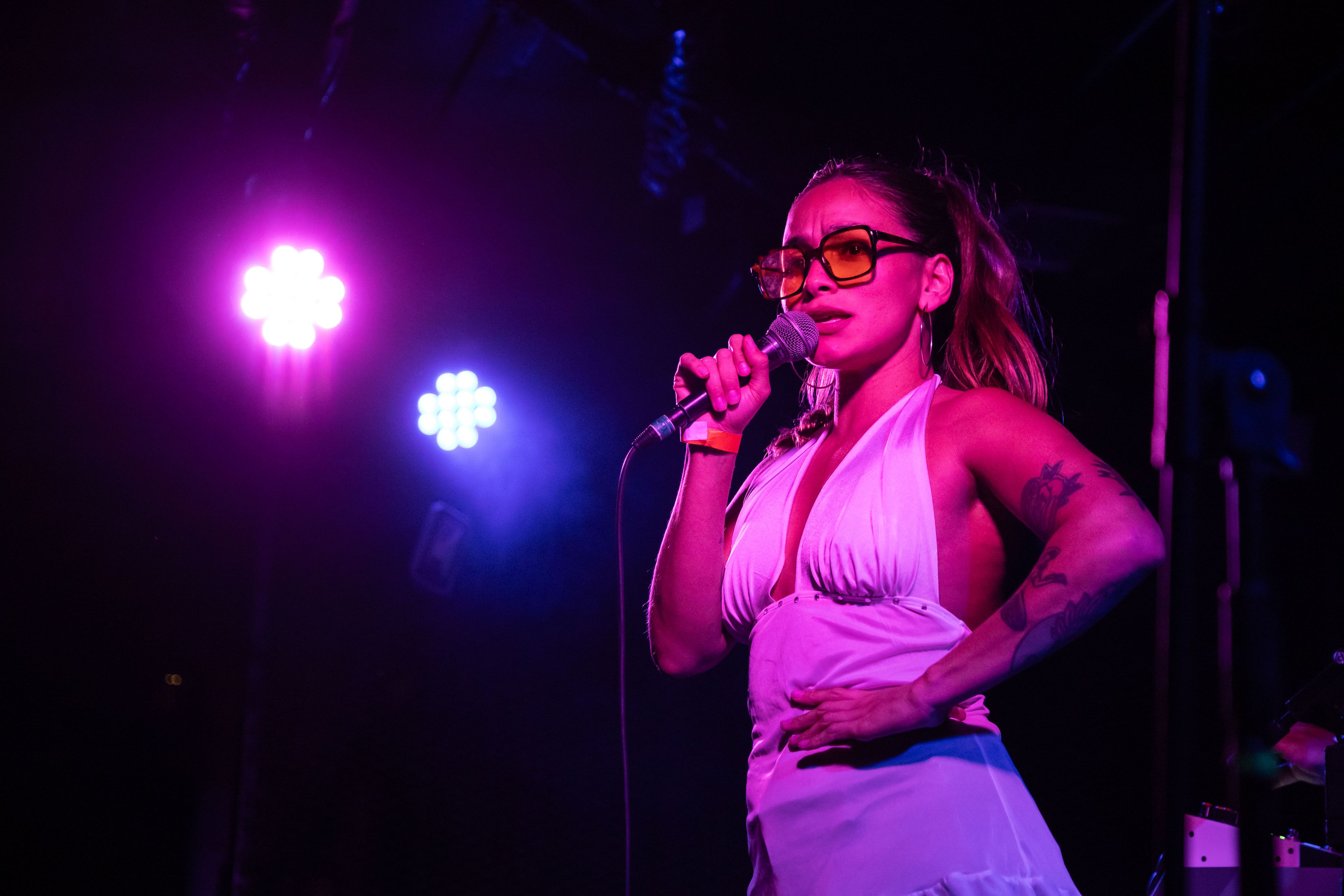 Michi stands with her hand on her hip singing into a microphone bathed in purple light