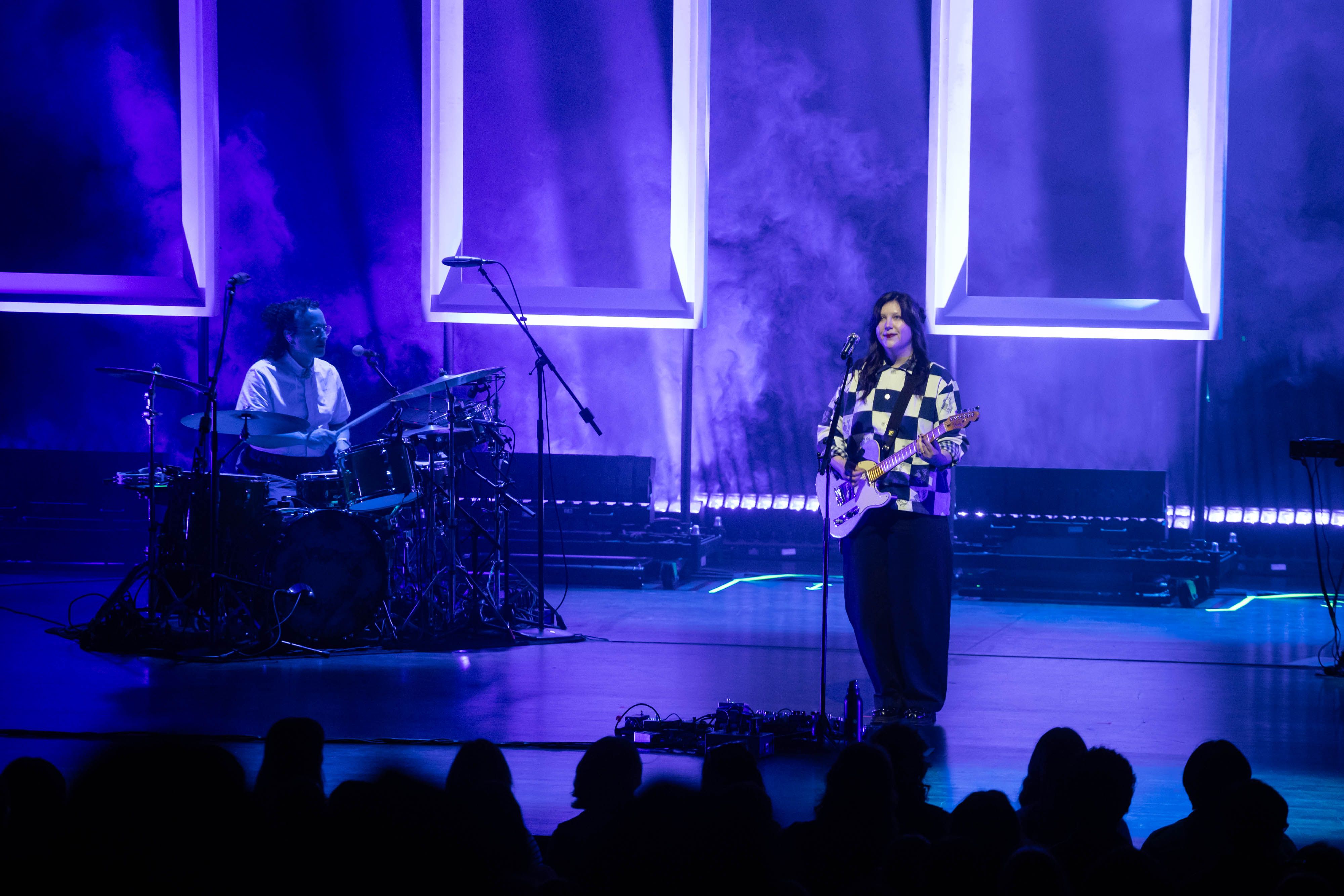 Lucy Dacus stands on the stage of the QE Theatre backlit by purple uplighting hitting empty frames behind her